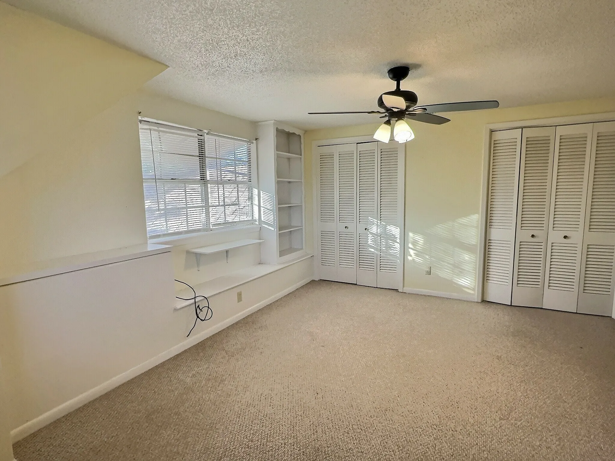 Carpeted room with a ceiling fan light fixture, two sets of louvered bi-fold closet doors, a built-in window bench, and integrated shelving