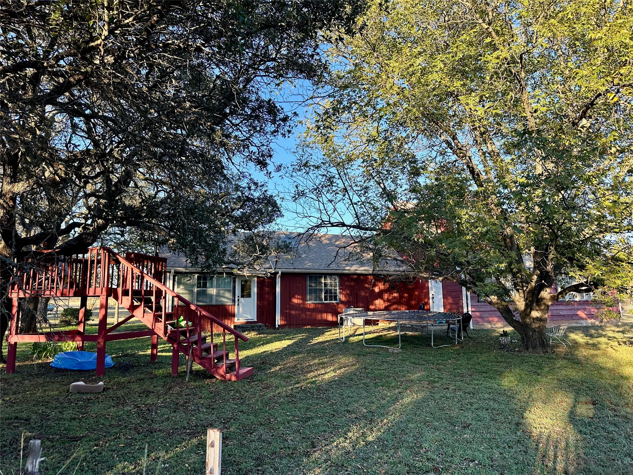 Expansive rear yard featuring mature trees, a multi-level wooden play structure, and a red-sided structure with multiple windows and a white entry door