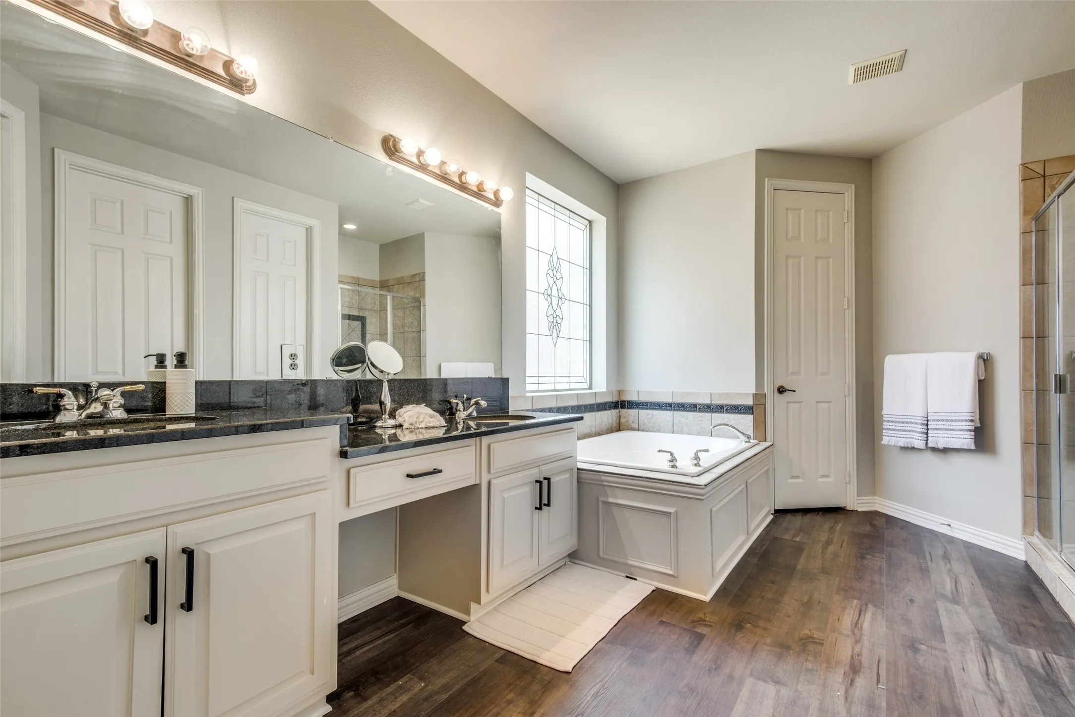 Bathroom featuring dual vanities with dark countertops, built-in soaking tub with decorative tile surround, a leaded glass window, and wood-finish flooring