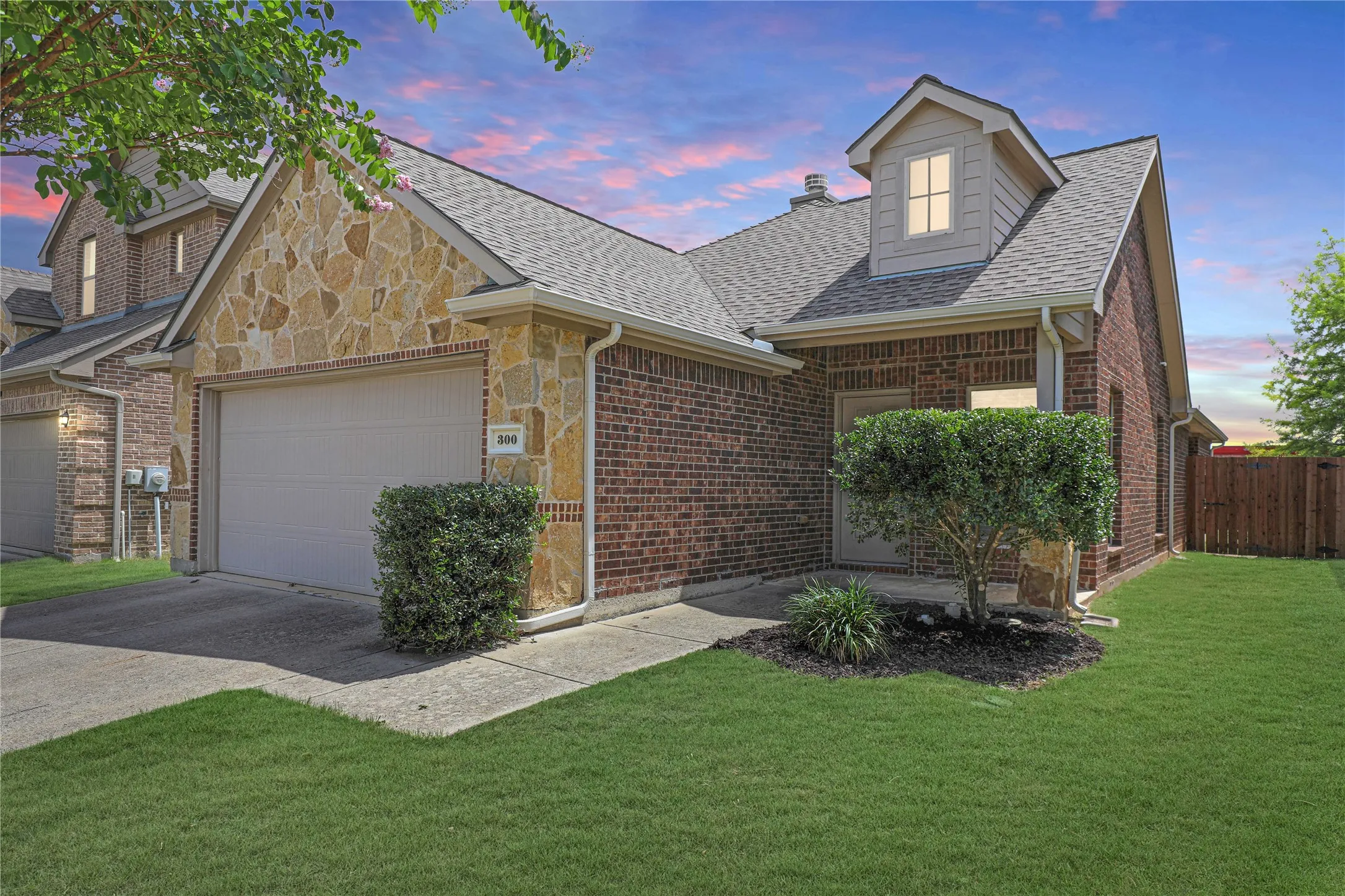 Brick and stone facade featuring a dormer window, attached garage, concrete driveway, and manicured landscaping
