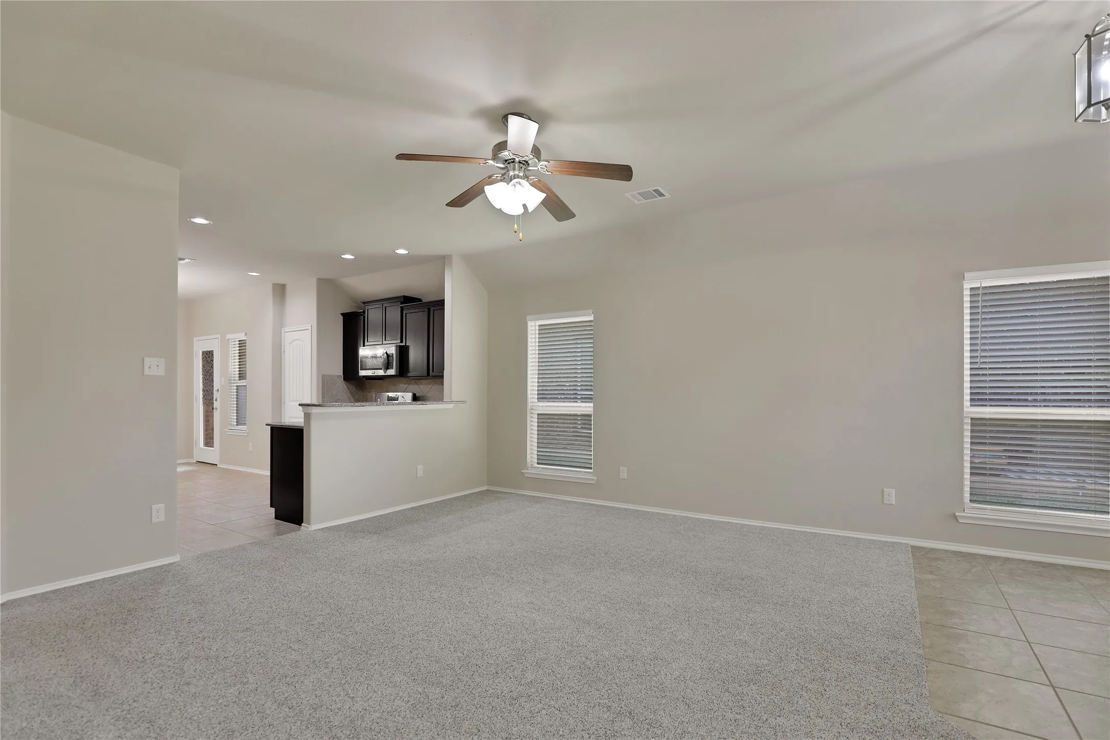 Spacious living area featuring grey carpet flooring, two windows with blinds, and a ceiling fan