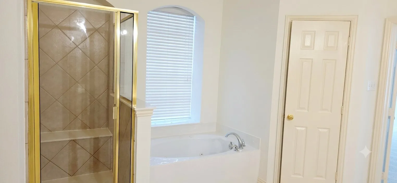 Tiled shower with gold-toned glass enclosure and built-in bench, accompanied by a soaking tub beneath an arched window with blinds