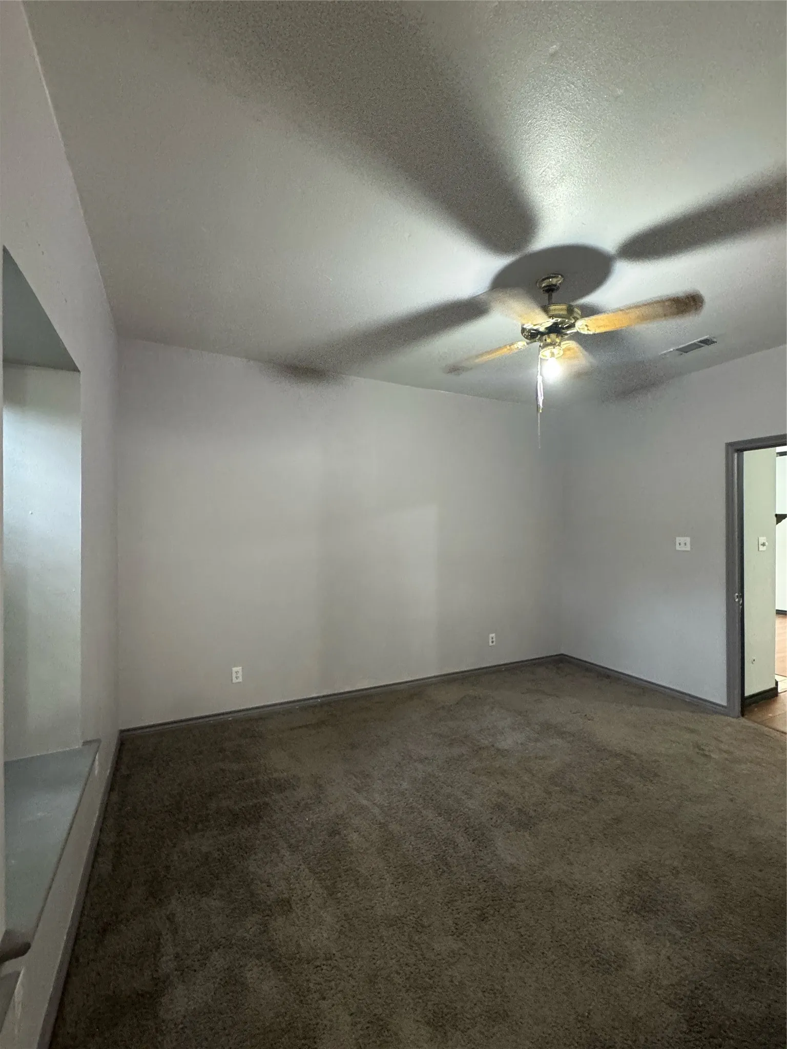Spacious interior room with neutral-toned walls, dark carpet flooring, and a ceiling fan with light fixture