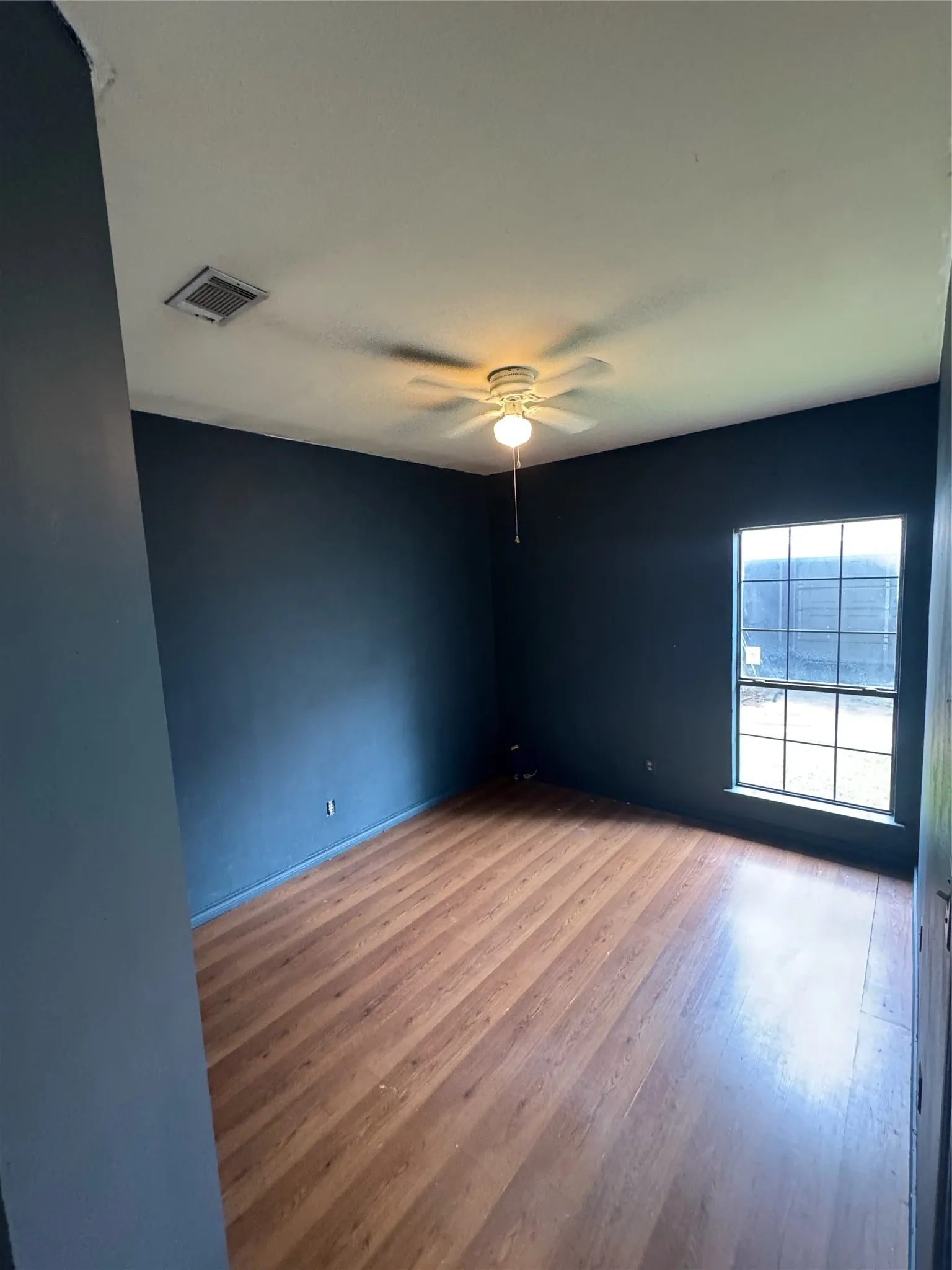 Spacious room featuring wood-finish flooring, a ceiling fan with integrated lighting, a large window with grilles, and dark-toned walls
