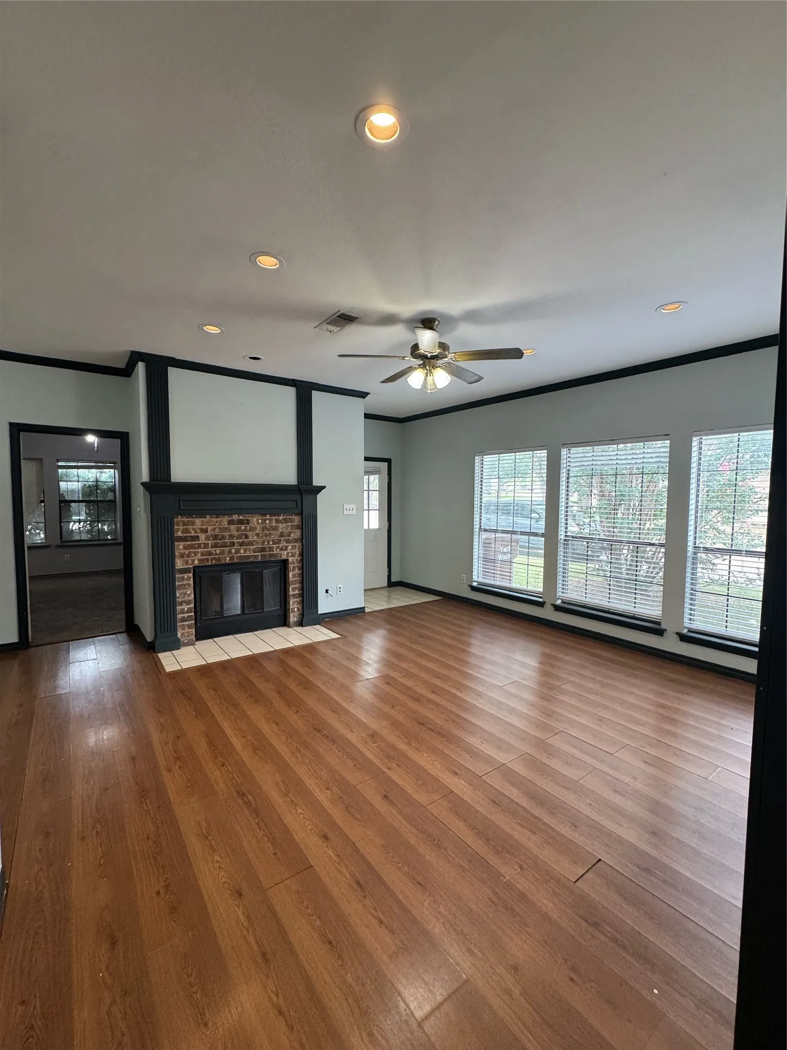 Spacious living area featuring wood-finish flooring, a brick fireplace with a mantel, and multiple windows with blinds