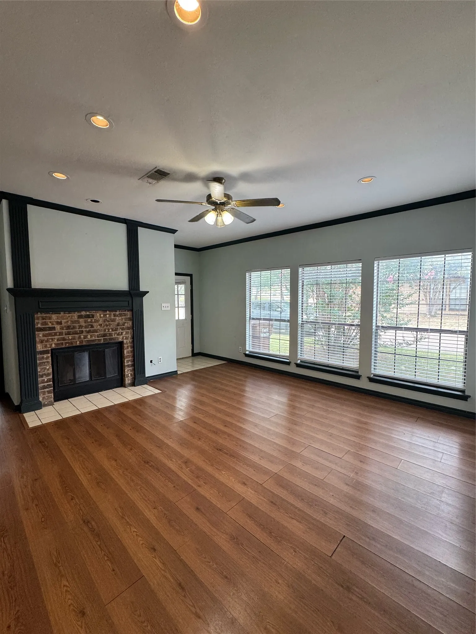Spacious living area featuring wood-finish flooring, a brick fireplace with a dark mantel, multiple windows with blinds, recessed lighting, and a ceiling fan