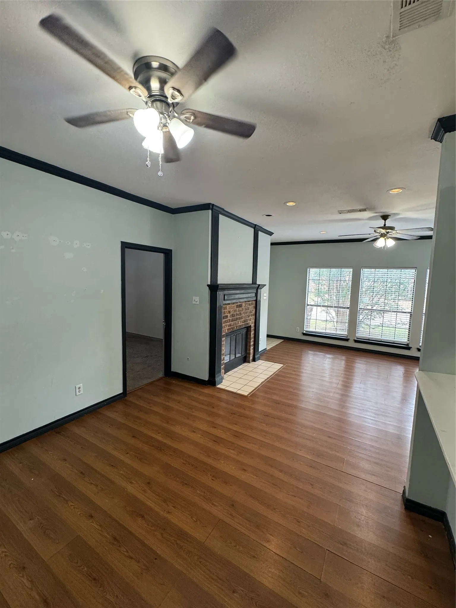 Spacious living area featuring wood-finish flooring, a brick fireplace with a tile hearth, and multiple ceiling fans