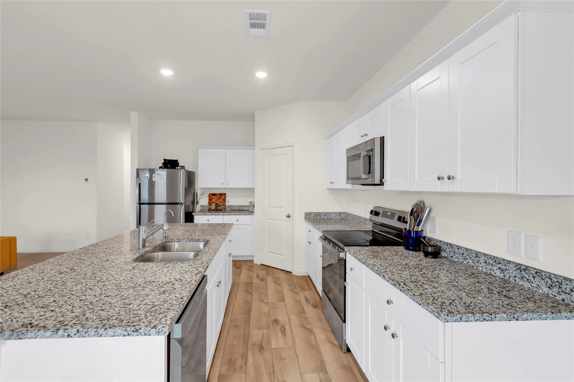 Modern kitchen featuring a spacious central island with a double basin sink, light-toned countertops, white cabinetry, stainless steel appliances, and wood-finish flooring