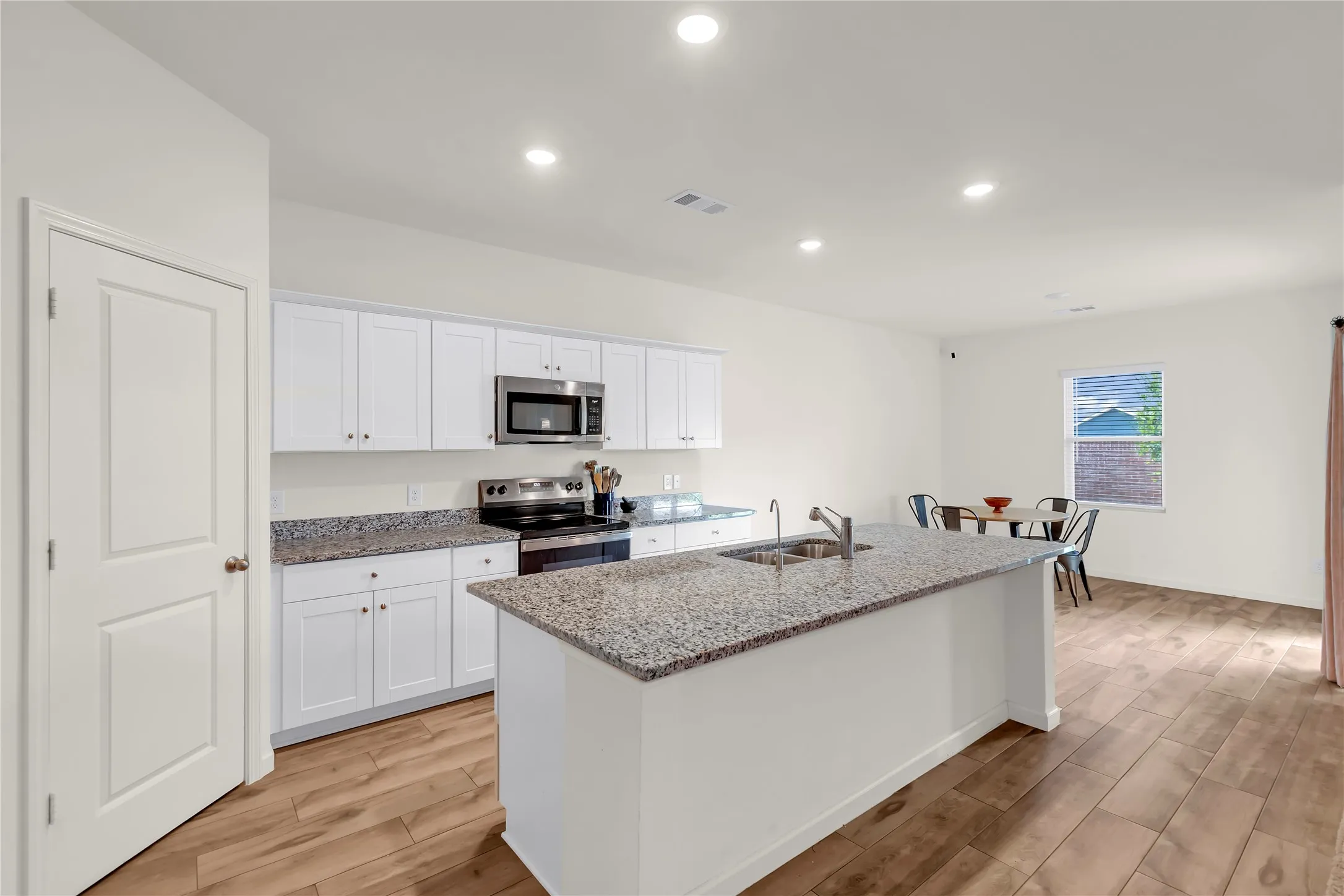 Open-concept kitchen featuring an island with an undermount sink, granite countertops, white cabinetry, stainless steel appliances, and wood-finish flooring
