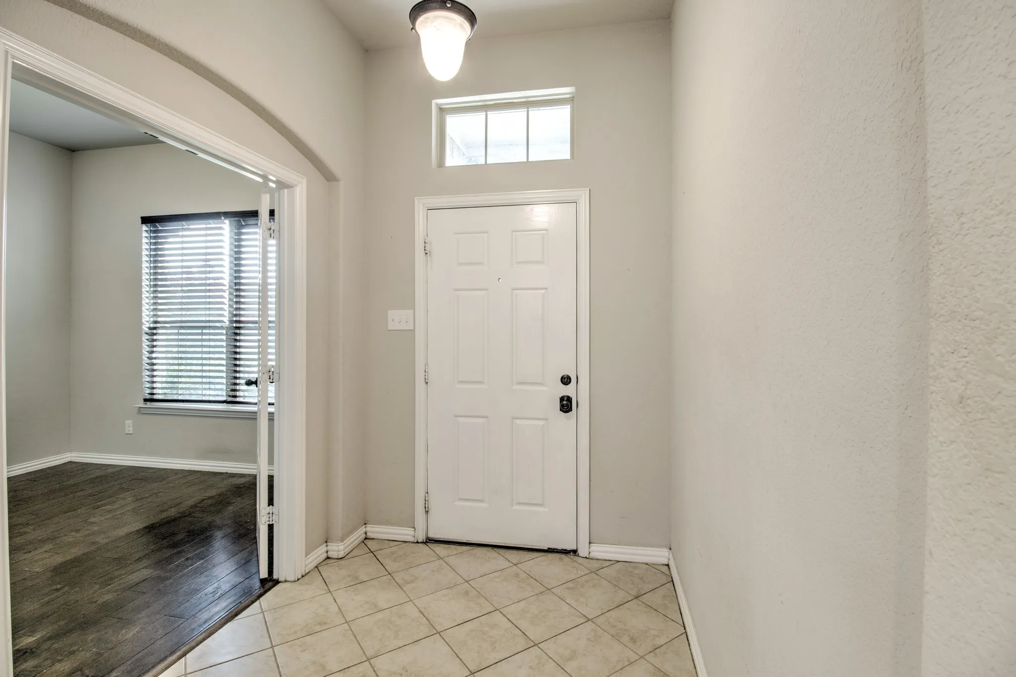 Foyer entry with neutral tile flooring, leading to a room featuring wood-finish flooring and a window with blinds