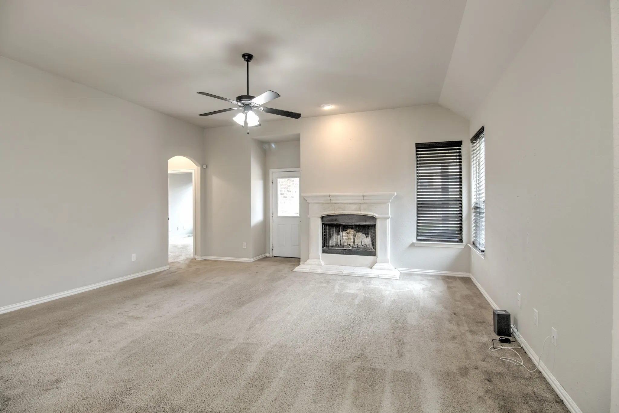 Spacious carpeted room featuring a fireplace with a light-colored mantel and hearth, vaulted ceiling, and an arched doorway