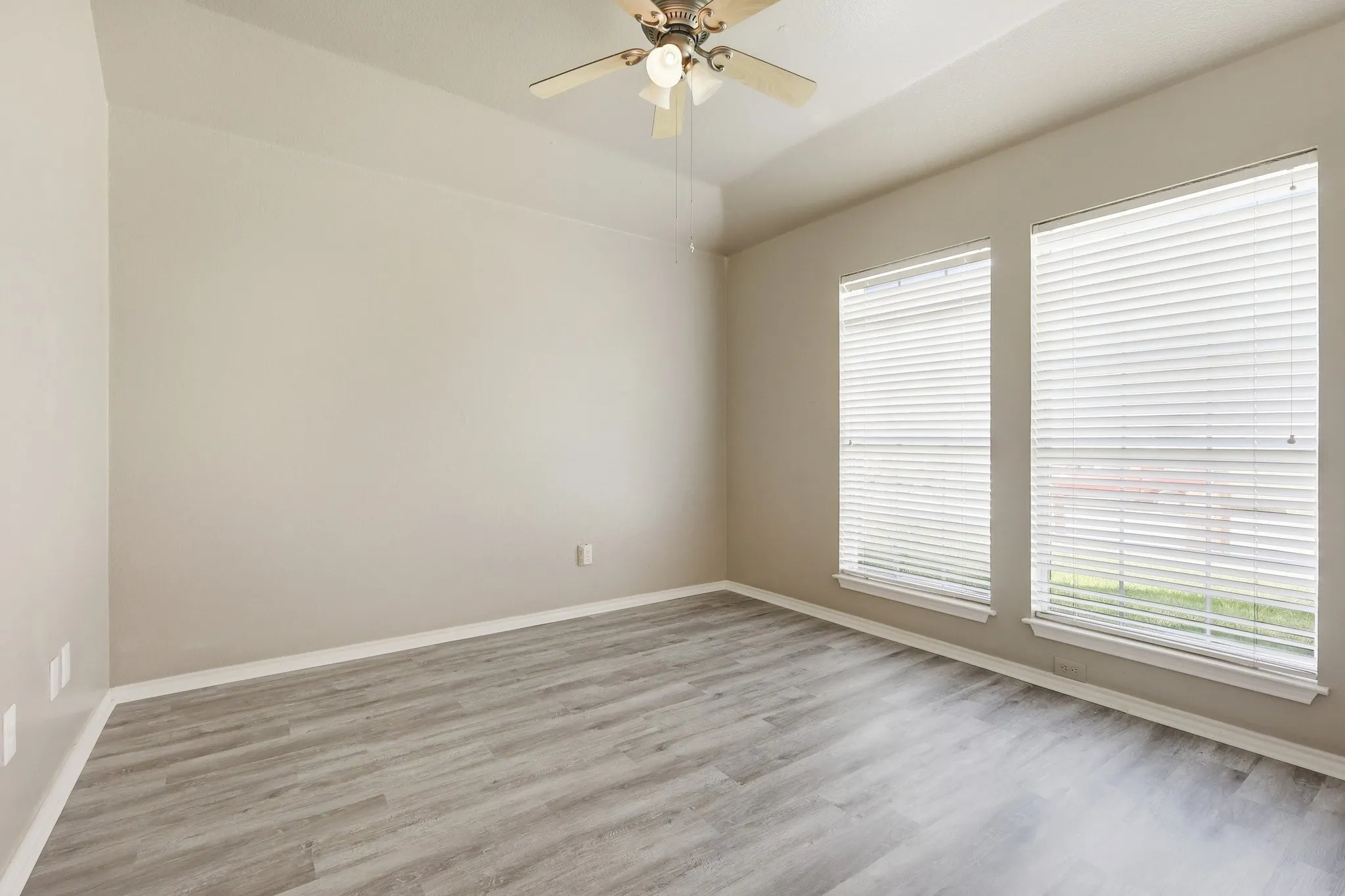 Spacious room featuring wood-finish flooring, two windows with blinds, a tray ceiling, and a ceiling fan with integrated lighting