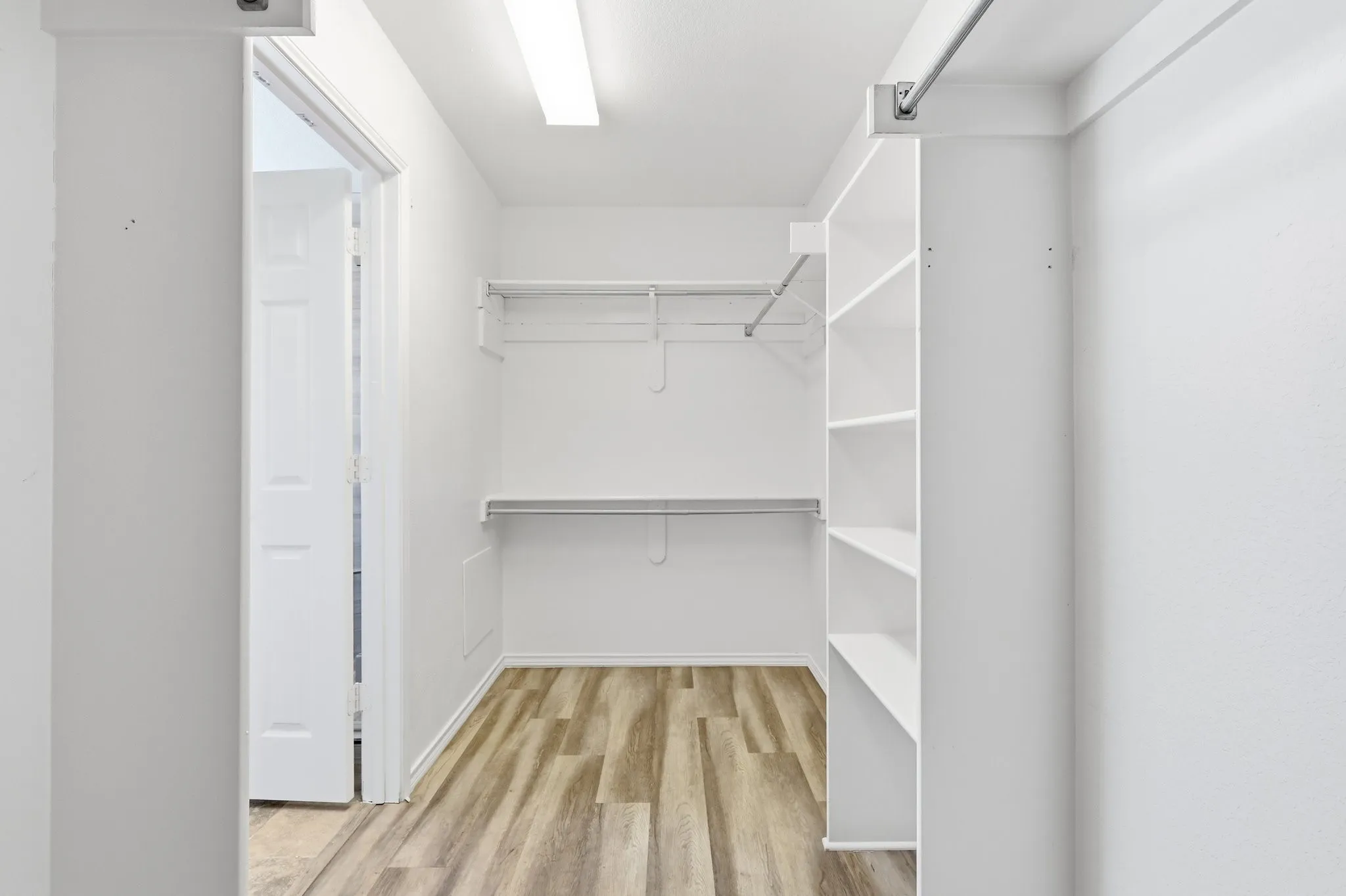 Walk-in closet featuring wood-finish flooring, built-in shelving, white walls, overhead fluorescent lighting, and metal hanging rods