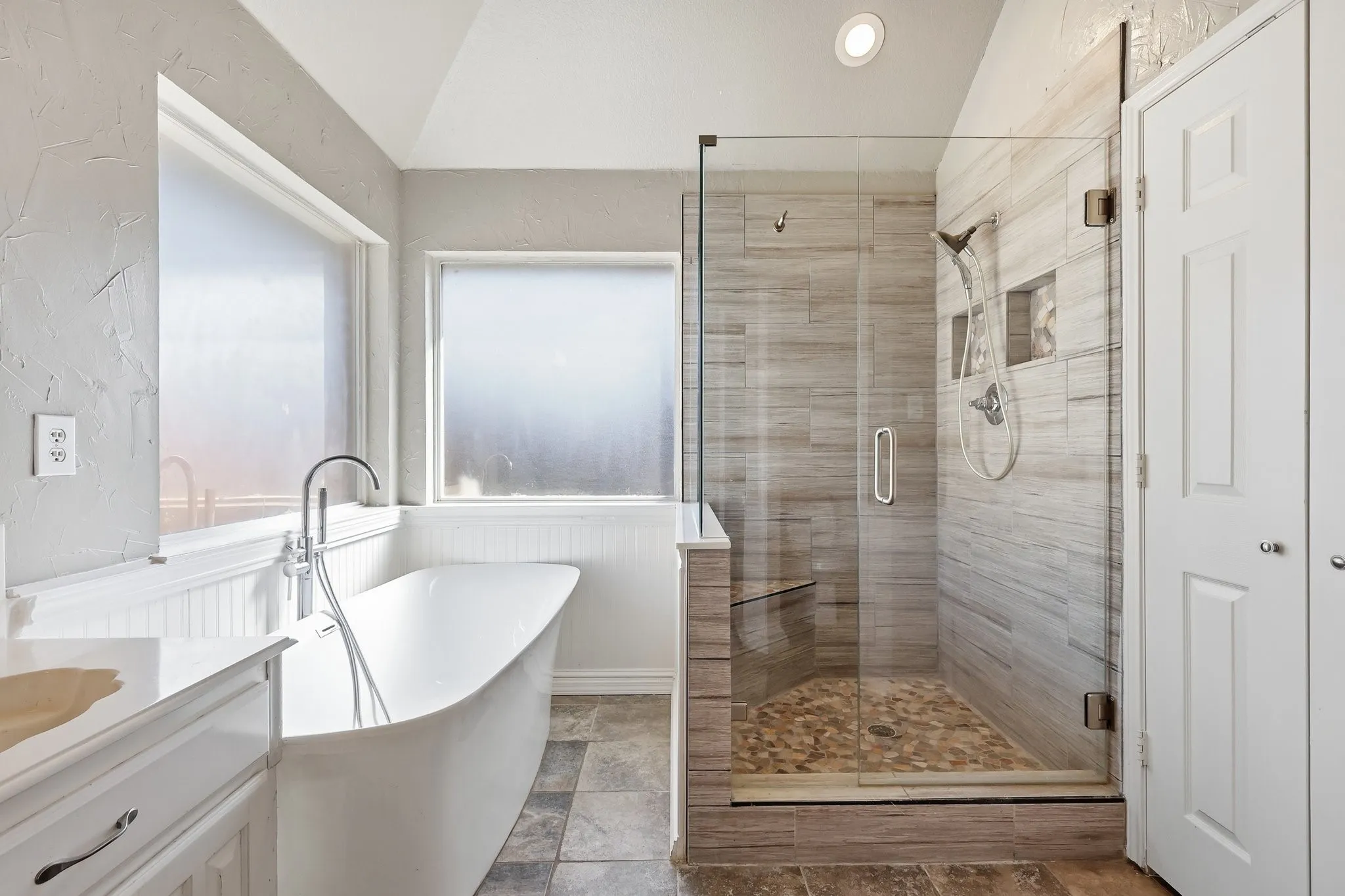 Freestanding soaking tub with a floor-mounted faucet, glass-enclosed shower with a pebble floor and tiled walls, and a white vanity with a light-toned countertop