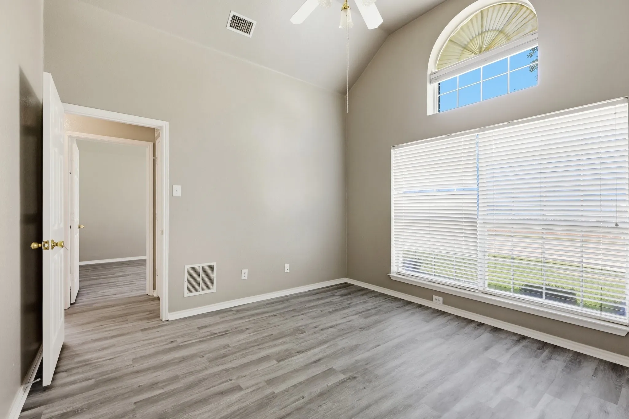 Vaulted ceiling room featuring light wood-finish flooring, a large window with blinds, an arched transom window, and a ceiling fan