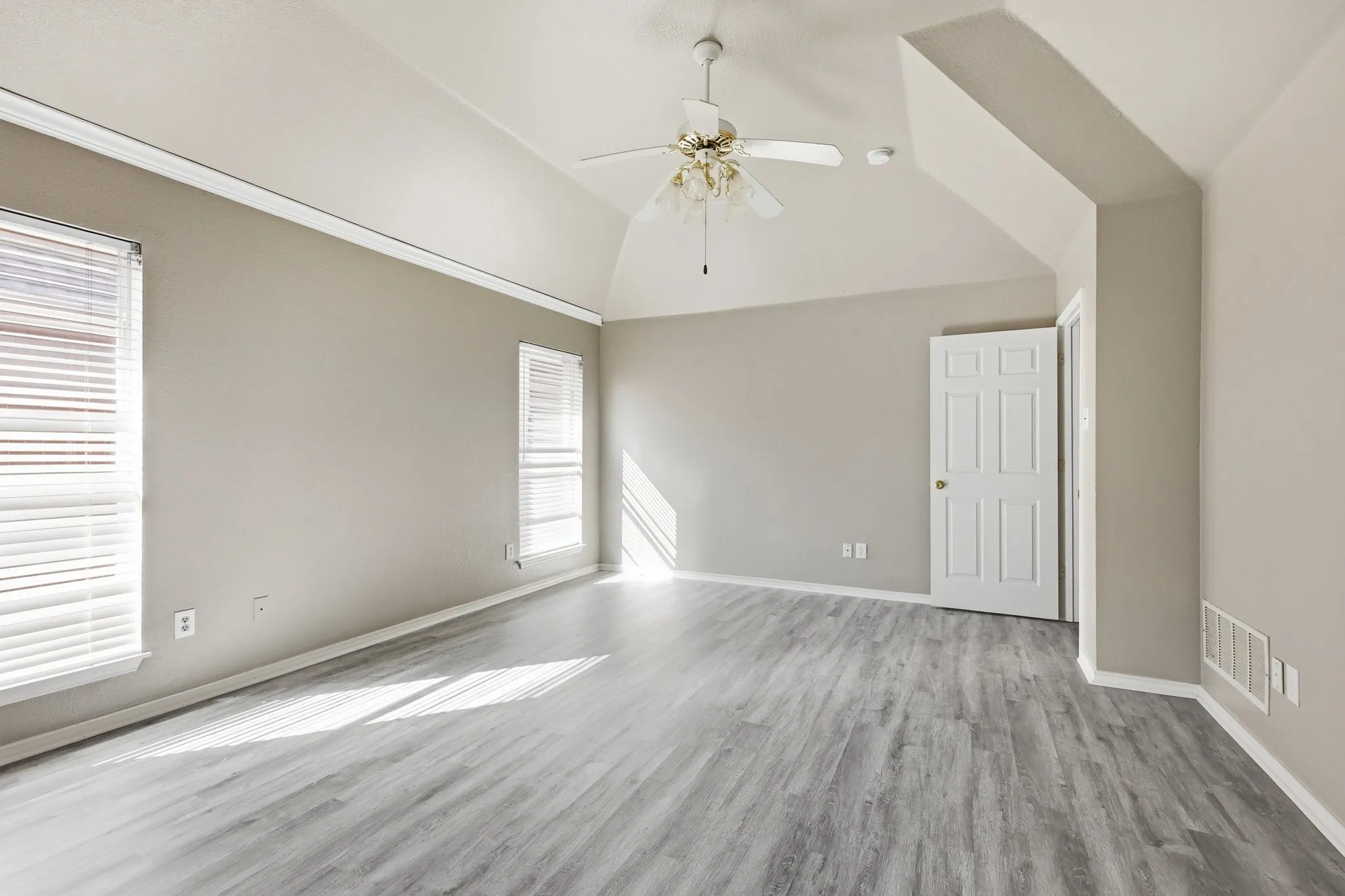 Spacious room featuring vaulted ceilings, a ceiling fan, two windows with blinds, wood-finish flooring, and a paneled interior door