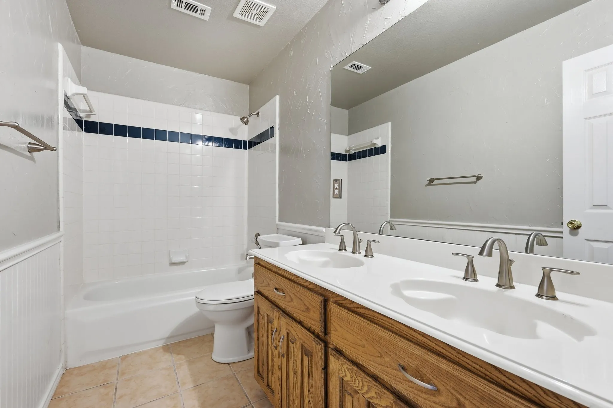 Bathroom featuring a double vanity with wood cabinetry and brushed nickel fixtures, a tub-shower combination with white subway tile and a dark accent band, and ceramic tile flooring