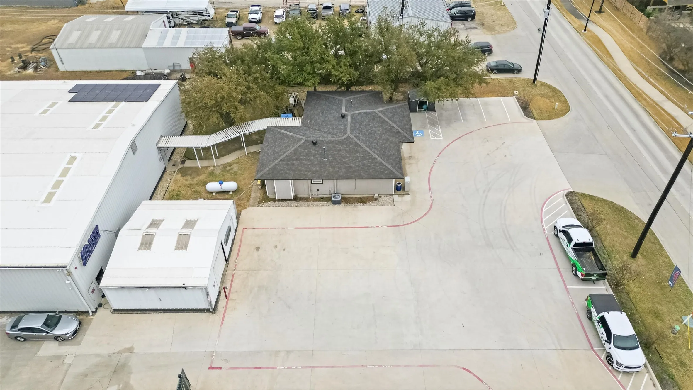 Commercial property featuring a gray shingle roof structure, expansive concrete parking areas, and adjacent large industrial buildings with solar panels