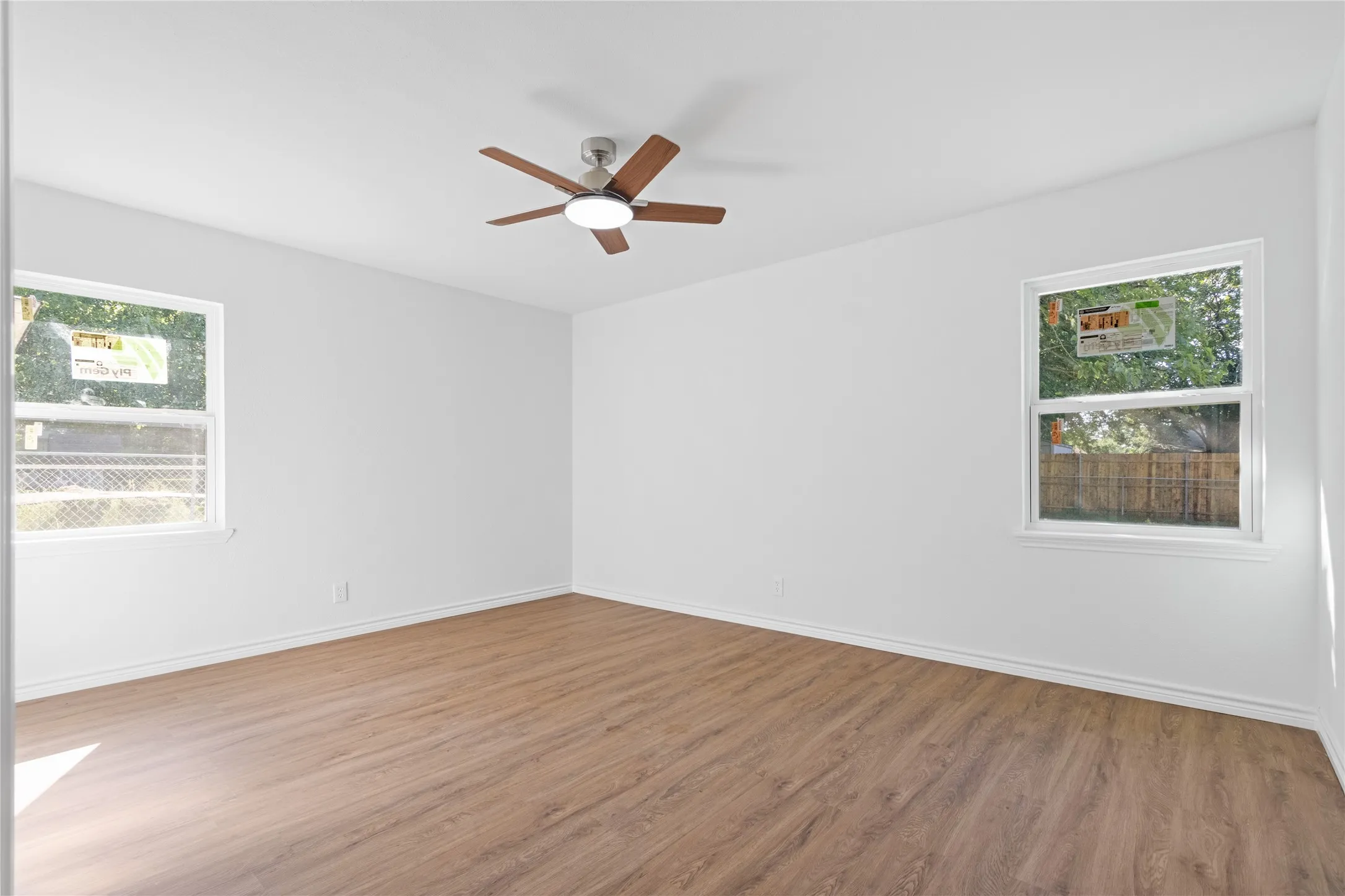 Spacious room featuring wood-finish flooring, a ceiling fan with integrated lighting, and two windows providing natural light