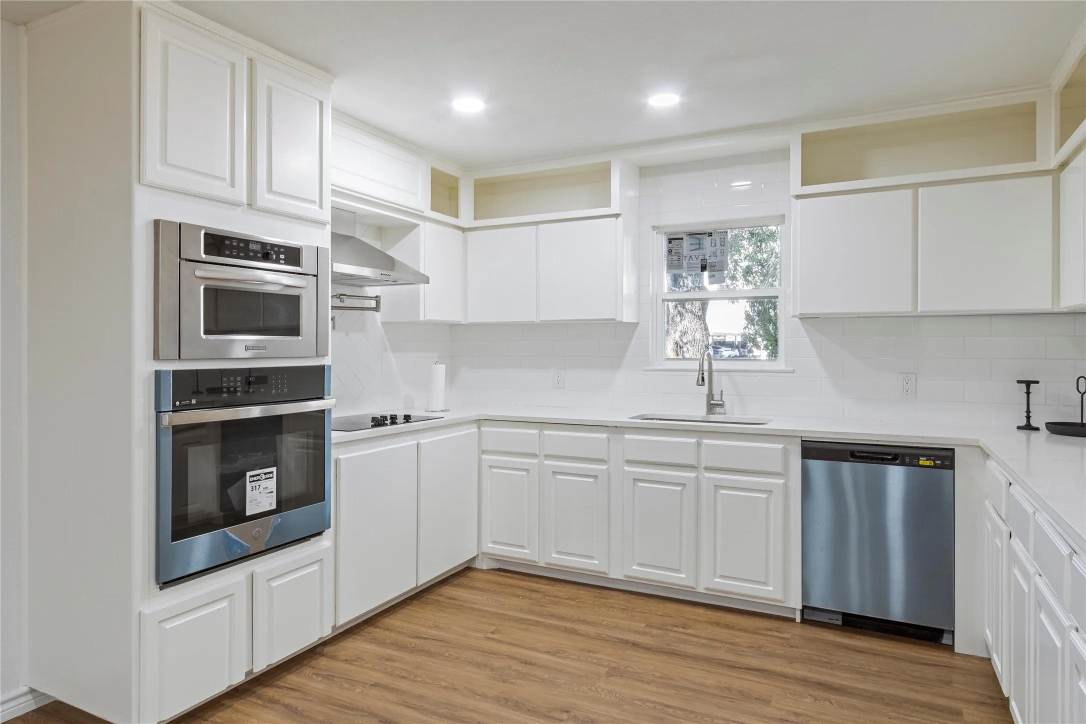 White shaker-style cabinetry, stainless steel appliances, white subway tile backsplash, light-toned countertops, and wood-finish flooring