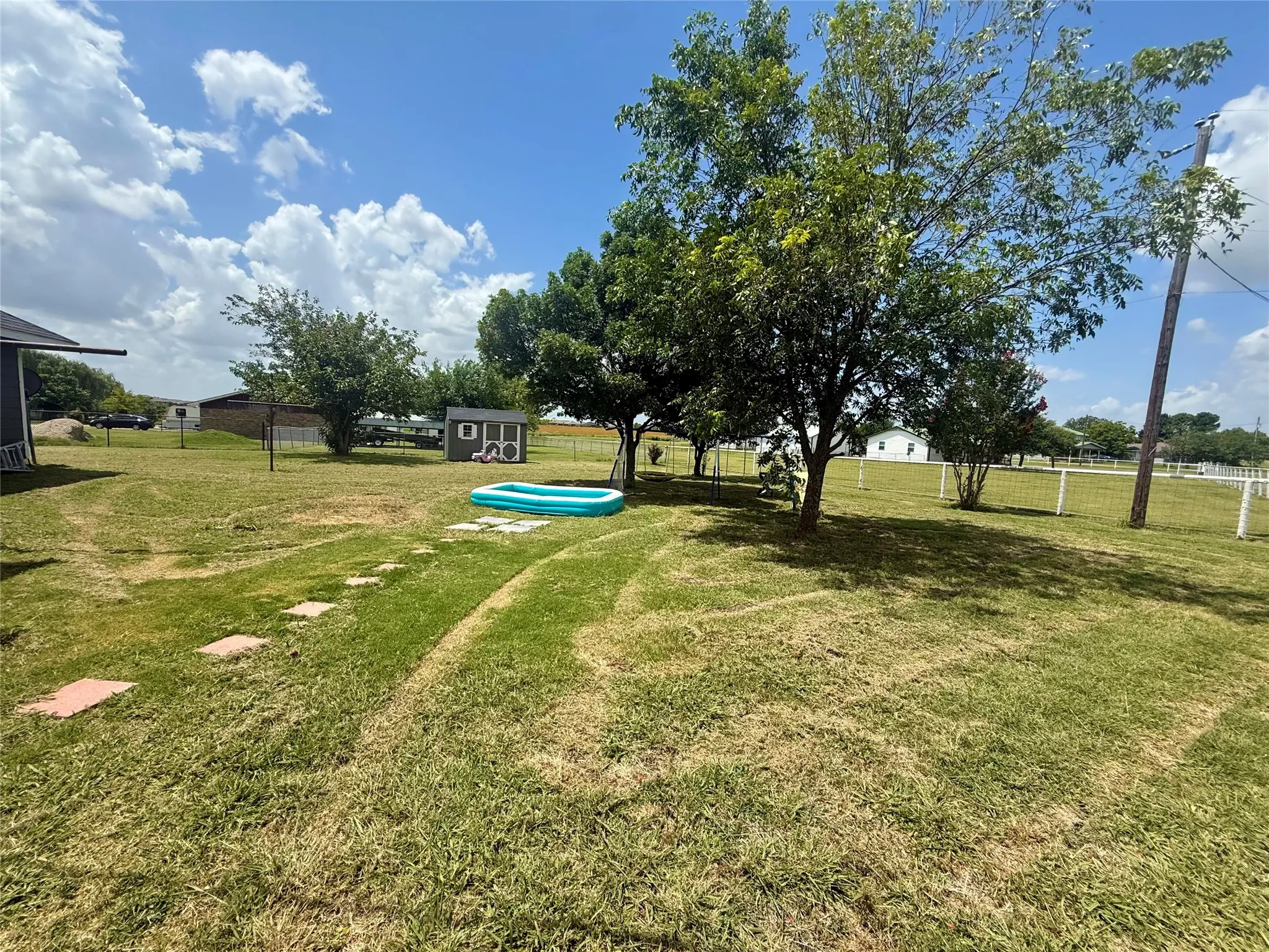 Expansive grassy yard featuring mature trees, a detached storage shed, stepping stones, and perimeter fencing