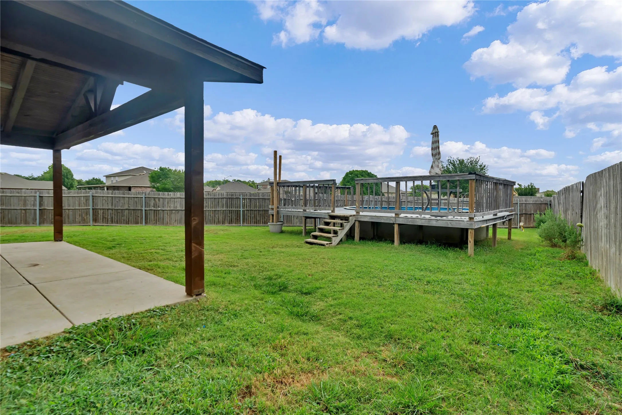 Covered patio and concrete walkway leading to an elevated outdoor pool with a surrounding wood deck, all within a fenced backyard