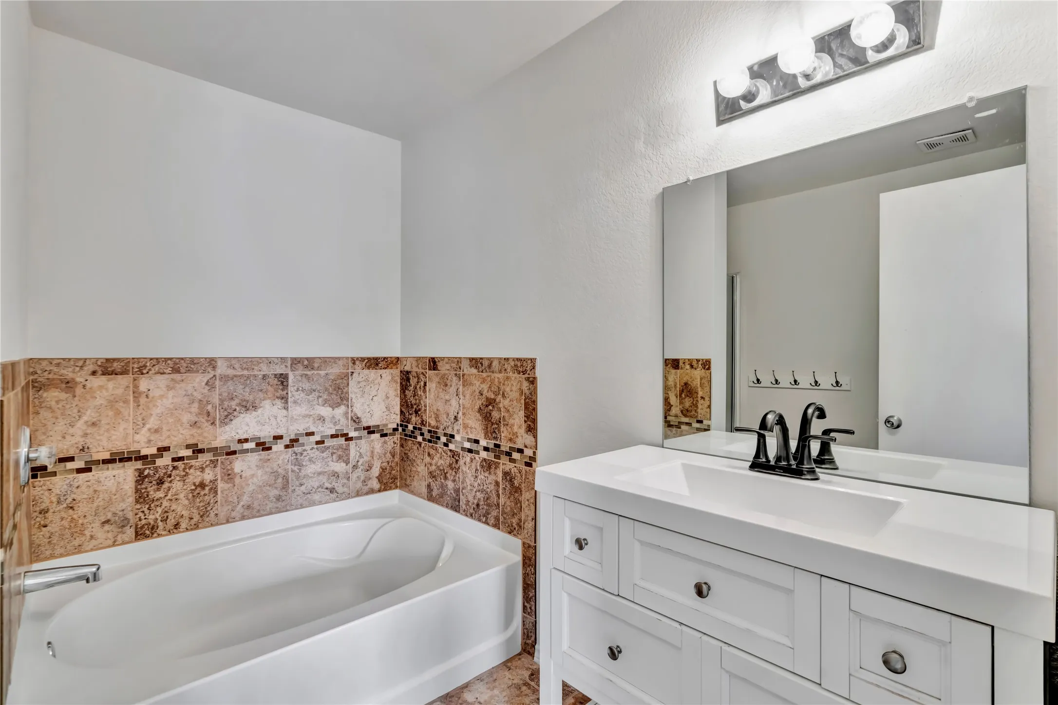 Bathroom featuring a white built-in tub, brown patterned tile surround with mosaic accent, white vanity with integrated sink, black faucet, and rectangular mirror with chrome light fixture