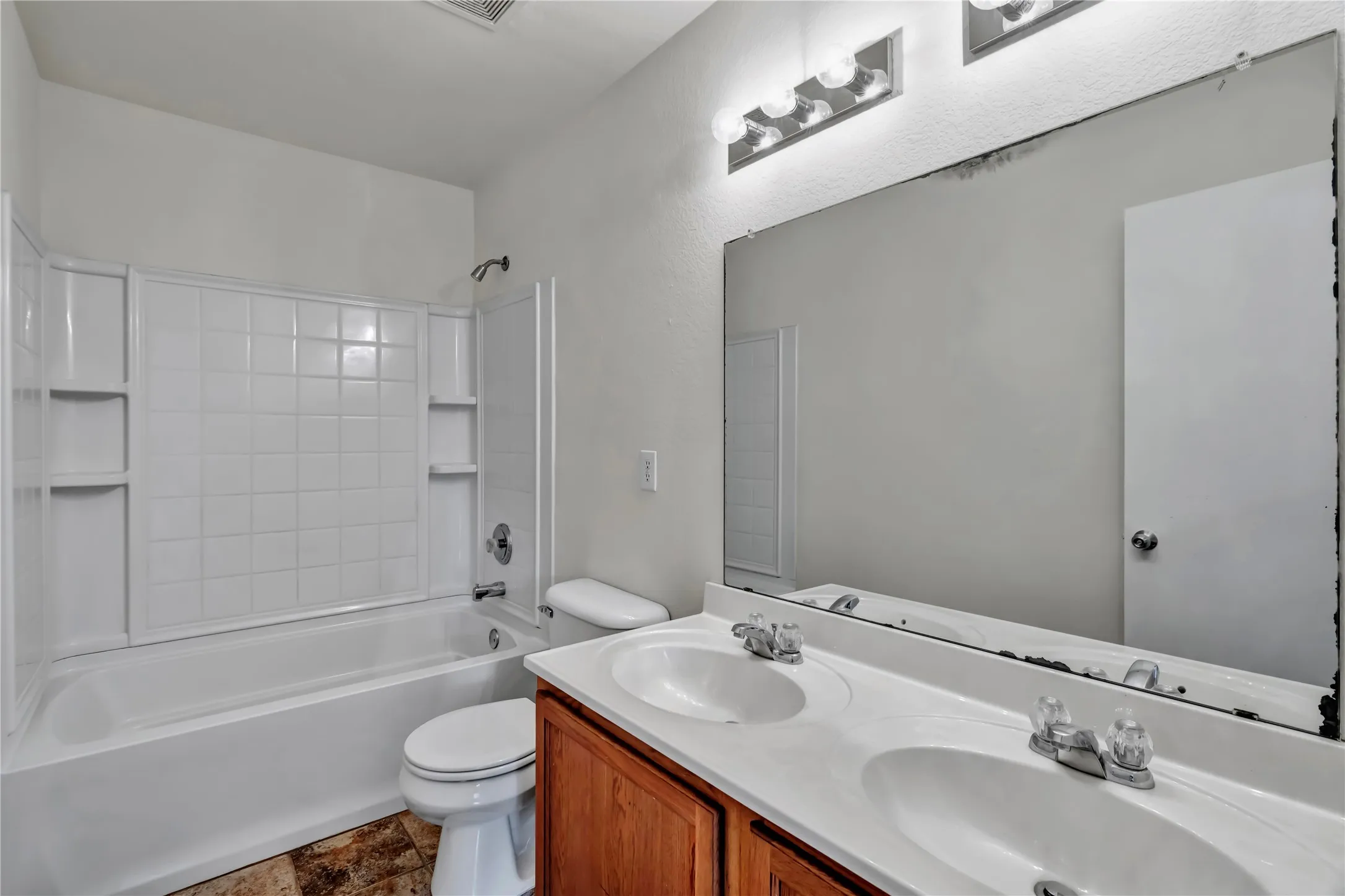 Bathroom featuring a dual-sink vanity with wood-finish cabinetry, a large wall-mounted mirror, overhead vanity lighting, and a bathtub with white tiled surround