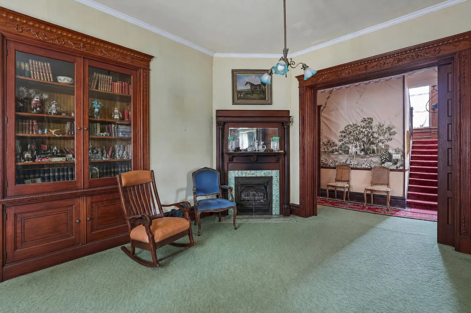 Room featuring a built-in wooden bookcase with glass-fronted doors, a fireplace with a wood mantle and tiled surround, and a wide wooden archway opening to a staircase with a red runner