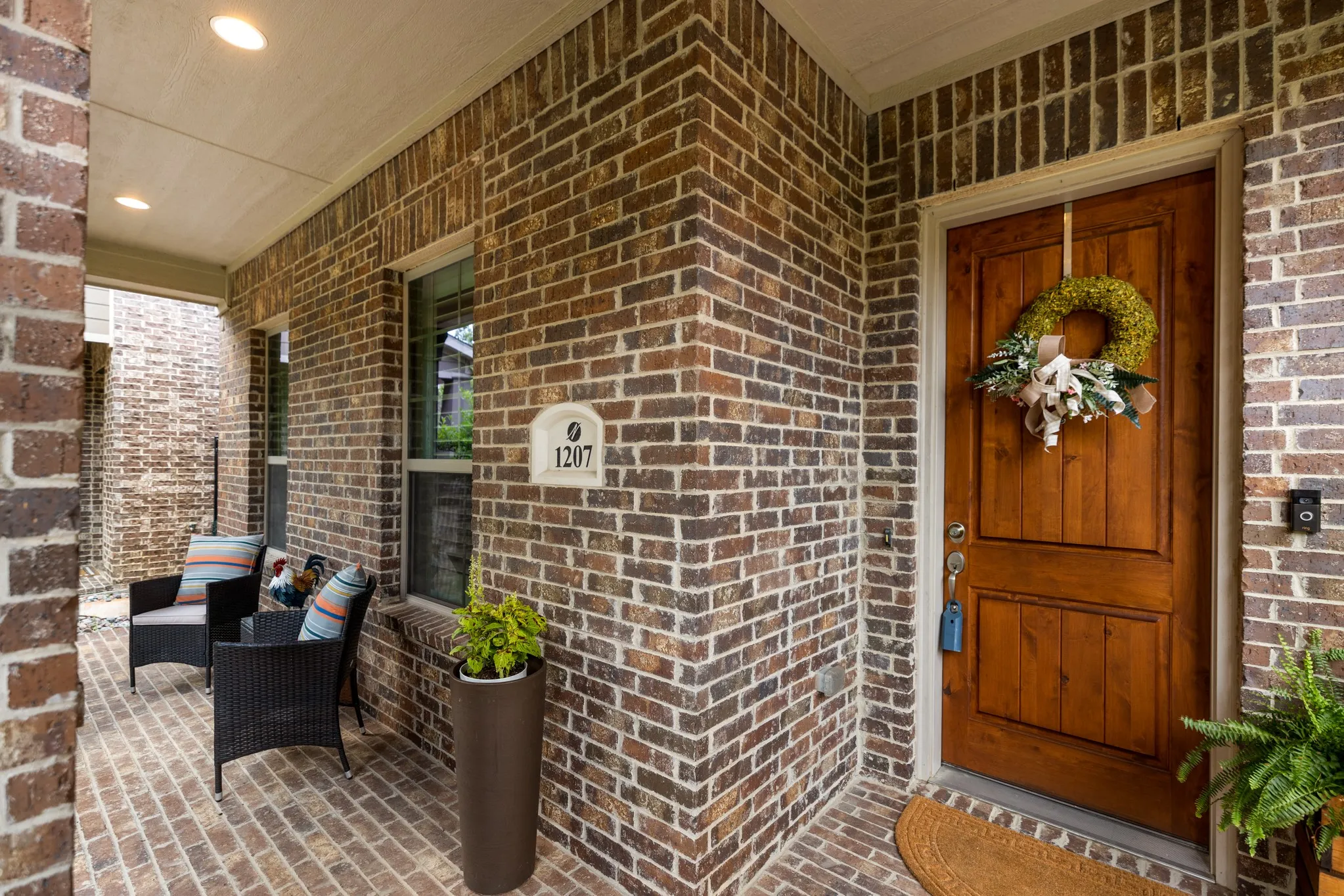 Covered front porch featuring a paneled wood-finish door, brick façade, recessed lighting, and brick pavers