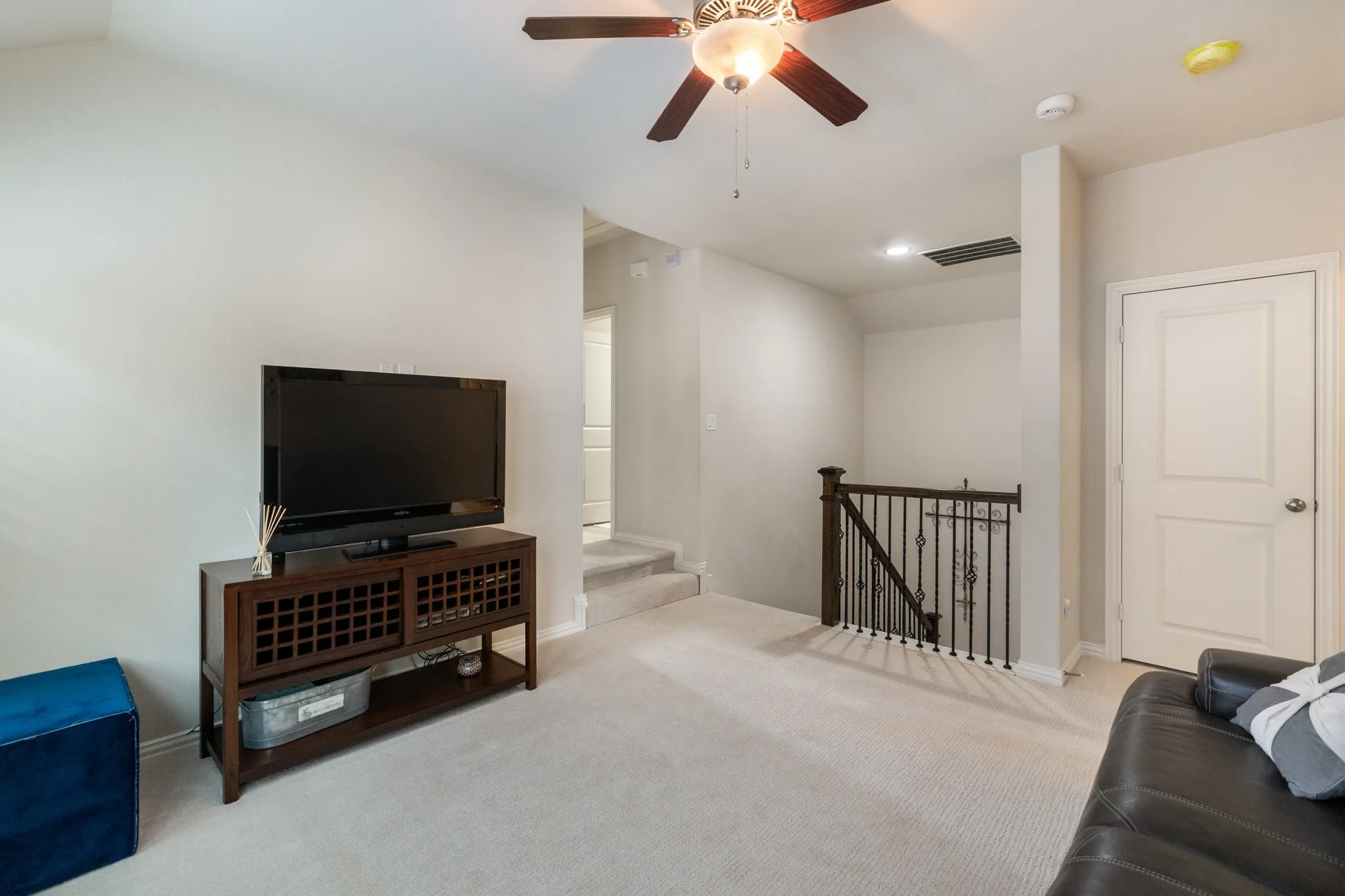 Carpeted room featuring a ceiling fan with light, white walls, and a staircase with wrought iron balusters and a dark wood handrail