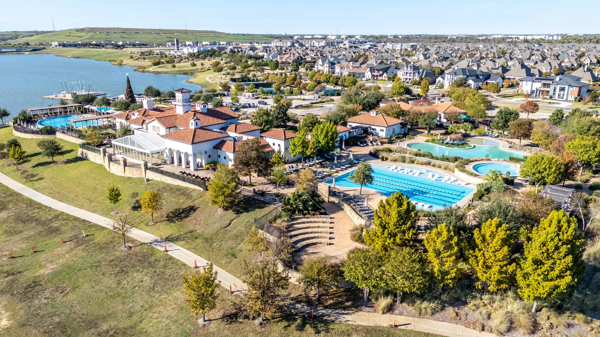 Spacious community grounds featuring multiple outdoor swimming pools, terraced amphitheater seating, a large clubhouse building with red tile roofing, and extensive paved walking paths
