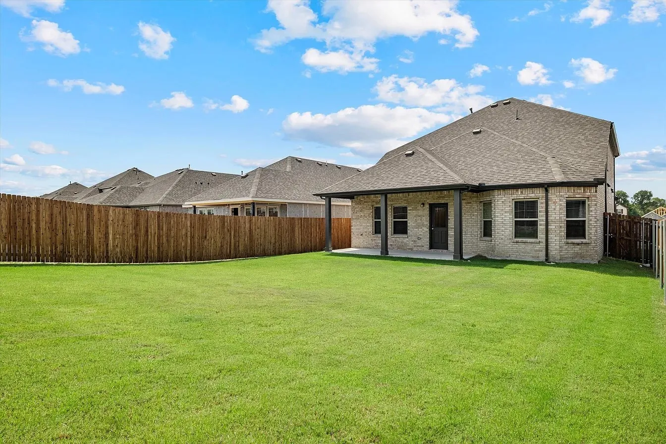 Expansive green lawn with a covered patio, rear brick exterior, asphalt shingle roof, and privacy fencing