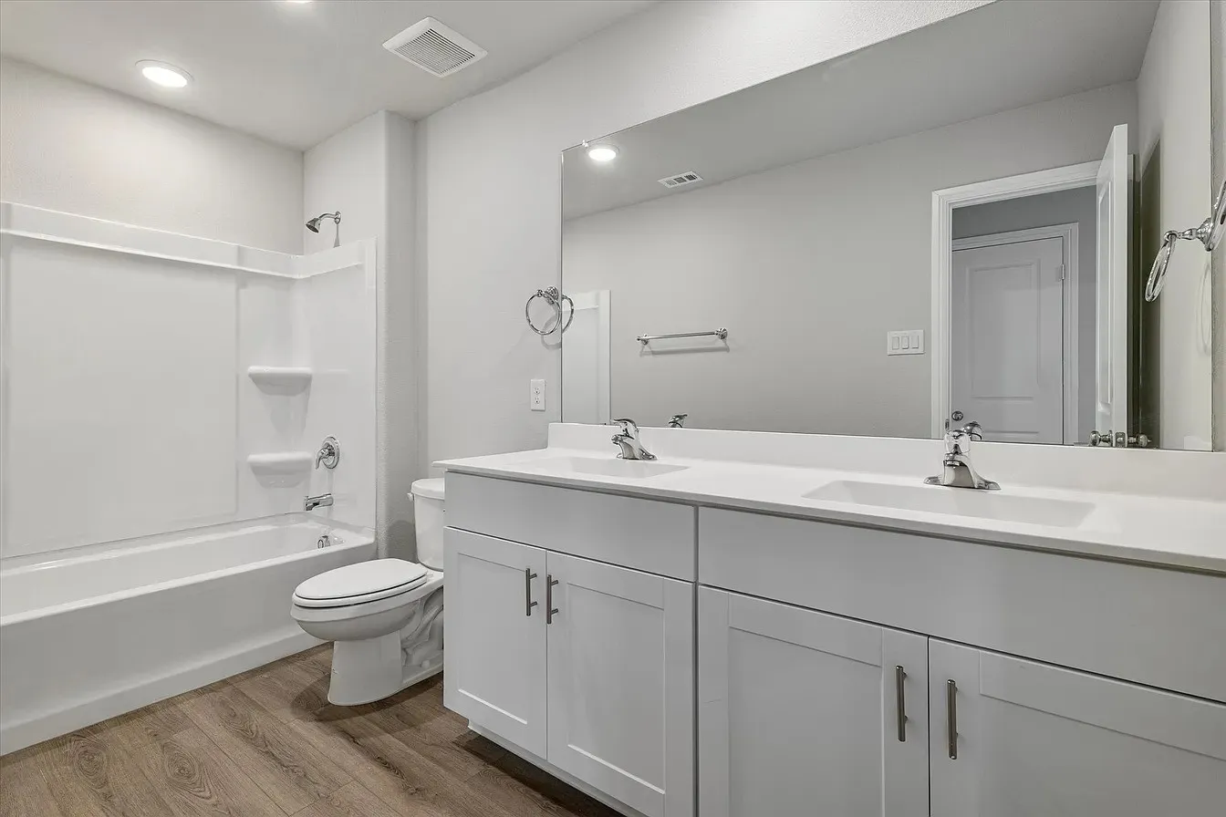 Bathroom featuring a dual vanity with white cabinetry and integrated sinks, a full-width wall mirror, a white bathtub with shower, and wood-finish flooring