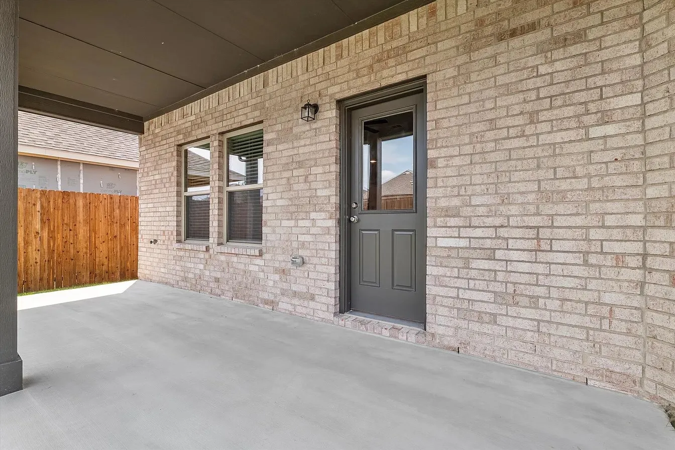 Covered outdoor patio featuring a concrete surface, light brick exterior, dark grey door with a paneled lower section and upper window, and two vertical windows with dark blinds