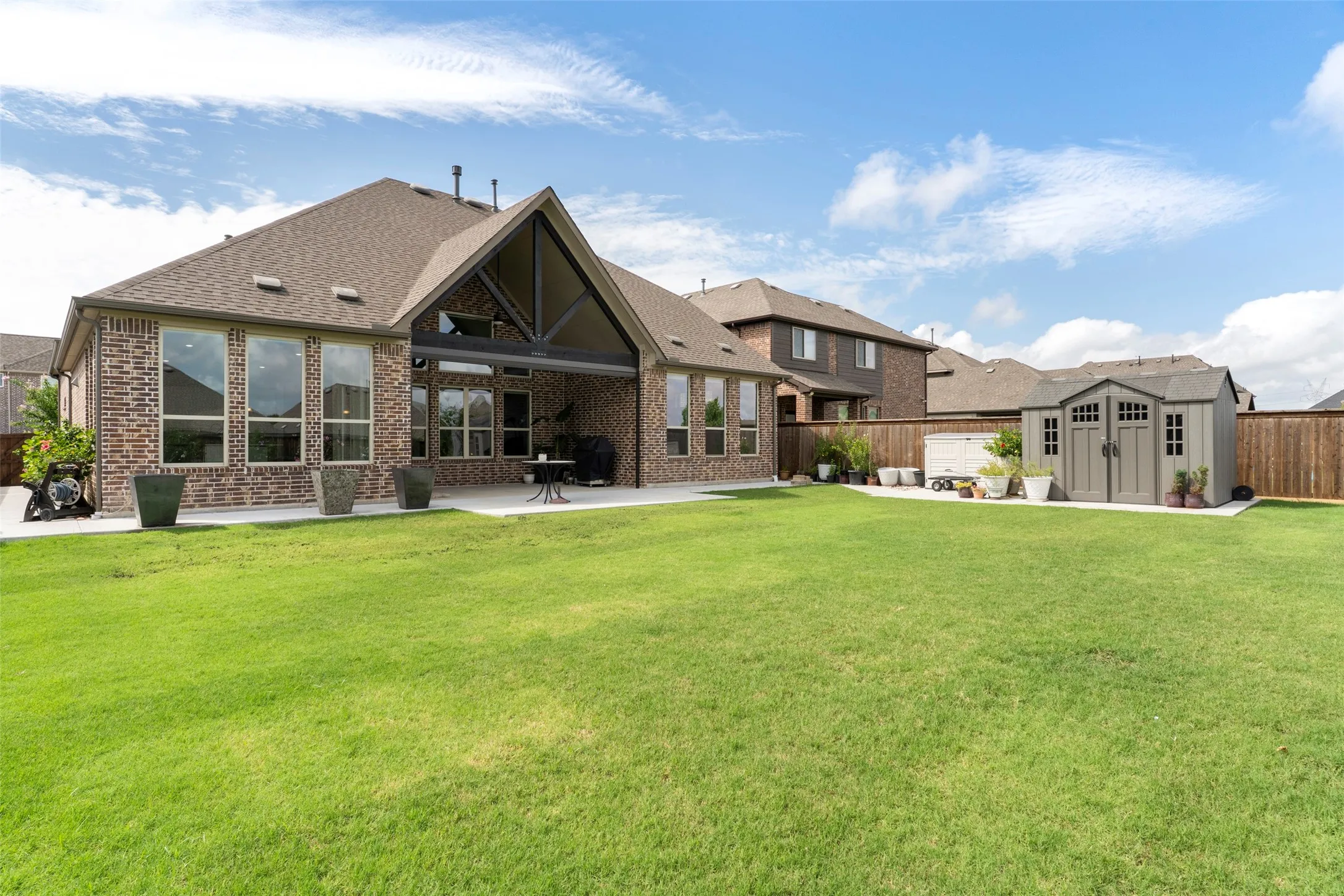 Expansive green lawn with a brick exterior home, featuring a covered patio, multiple large windows, and a detached storage shed