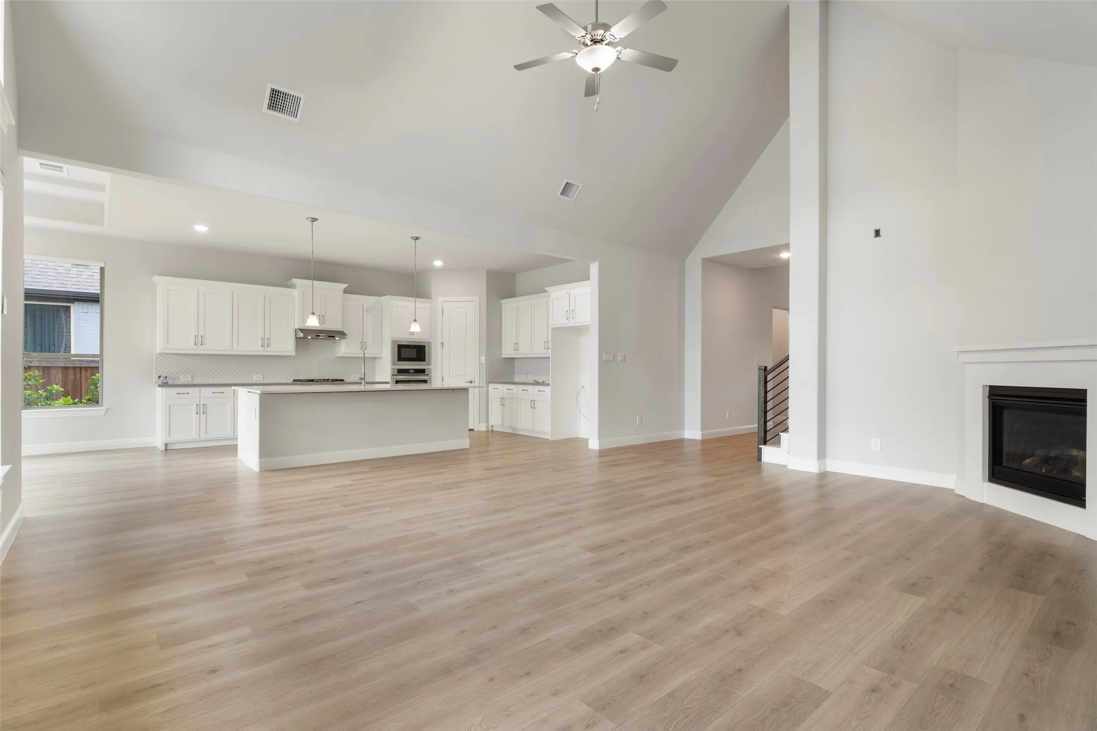Open-concept living area featuring light wood-finish flooring, a contemporary fireplace with white surround, vaulted ceilings, and an integrated kitchen with white cabinetry and a central island
