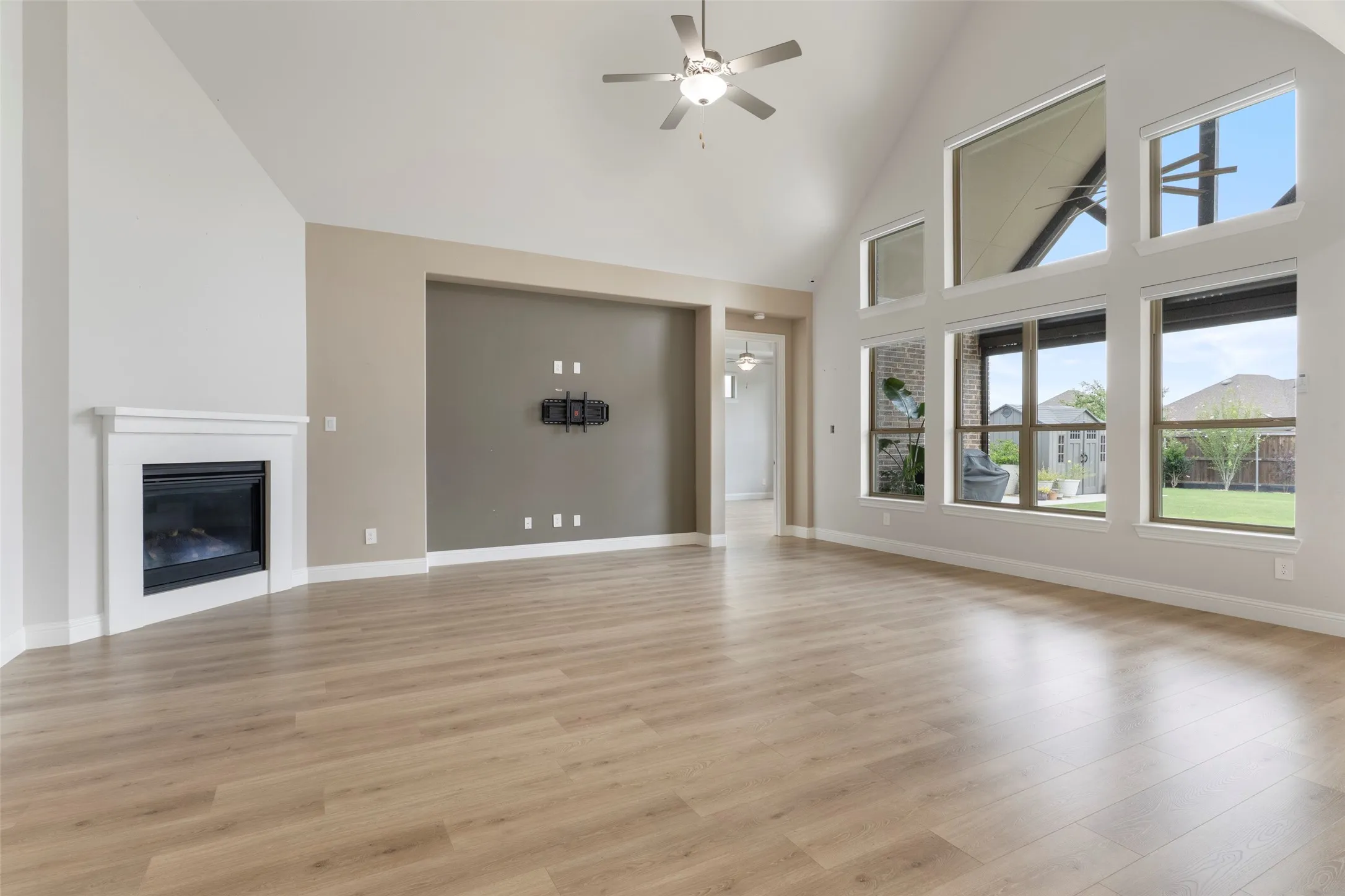 Spacious living area featuring vaulted ceilings, wood-finish flooring, a contemporary fireplace with white mantel, a ceiling fan with integrated lighting, and multiple large windows