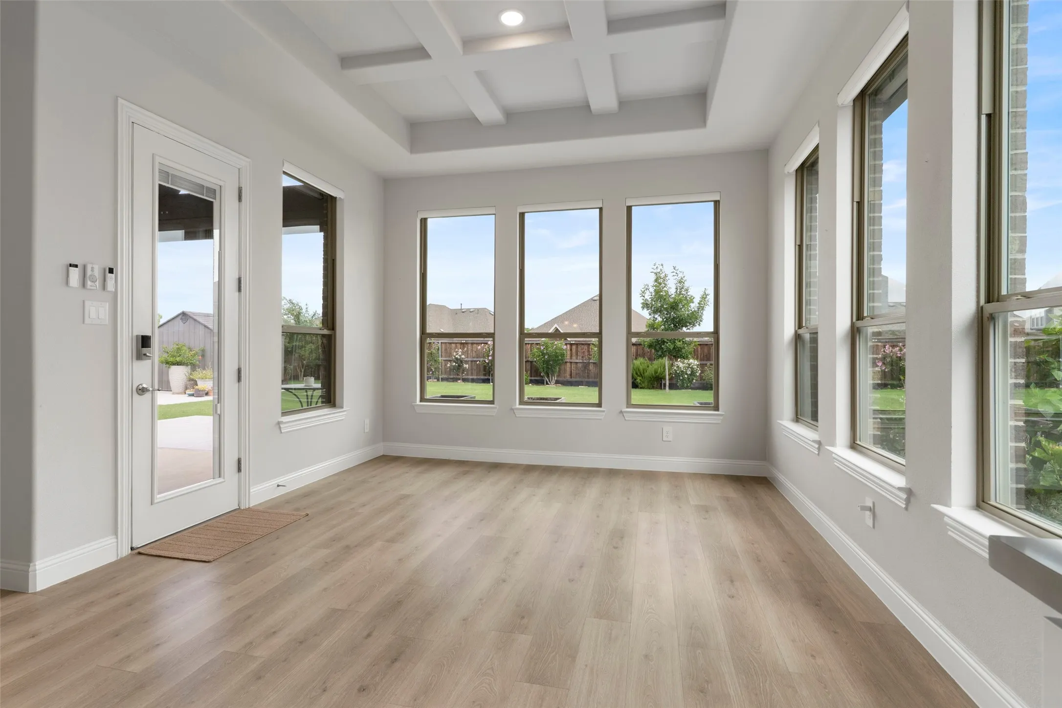 Spacious room featuring wood-finish flooring, a coffered ceiling, multiple large windows, a full-lite exterior door, and light gray wall paint with white trim