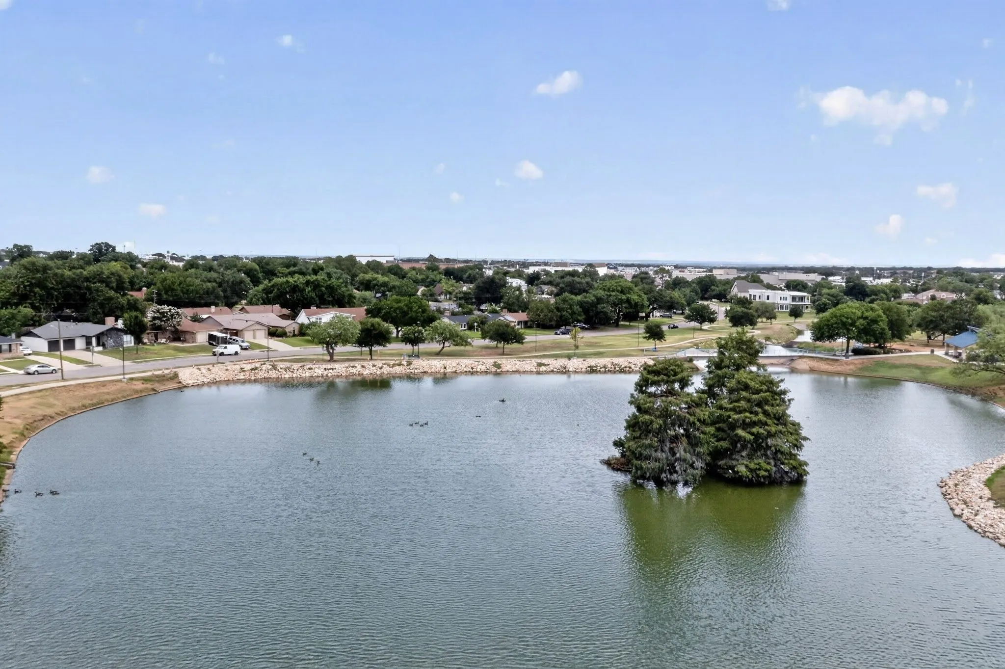 Expansive lake with a central tree-covered island, surrounded by a landscaped shoreline featuring rock accents and mature trees