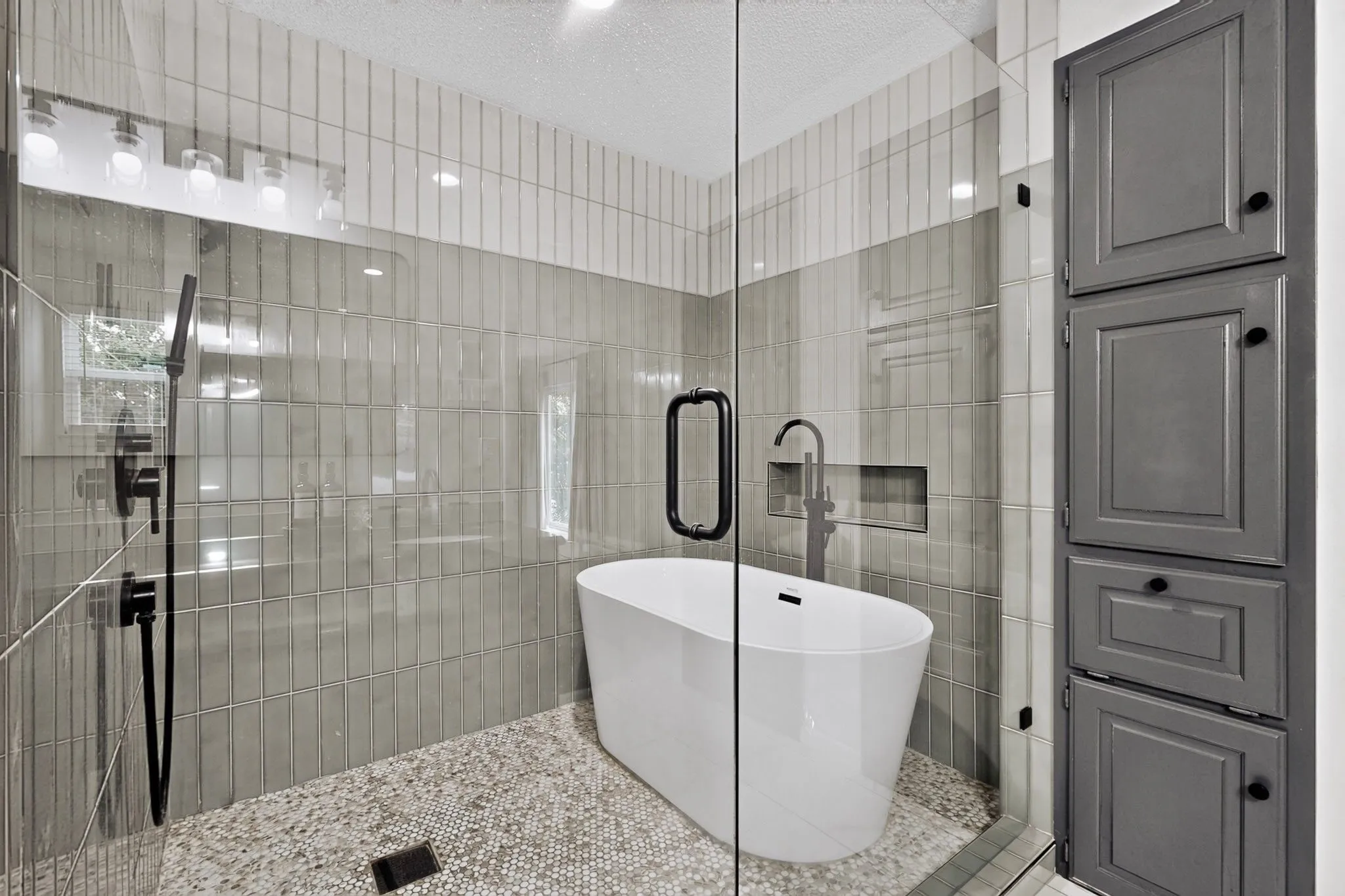 Glass-enclosed wet room featuring a freestanding soaking tub, mosaic tile flooring, vertical subway tile walls, and matte black fixtures