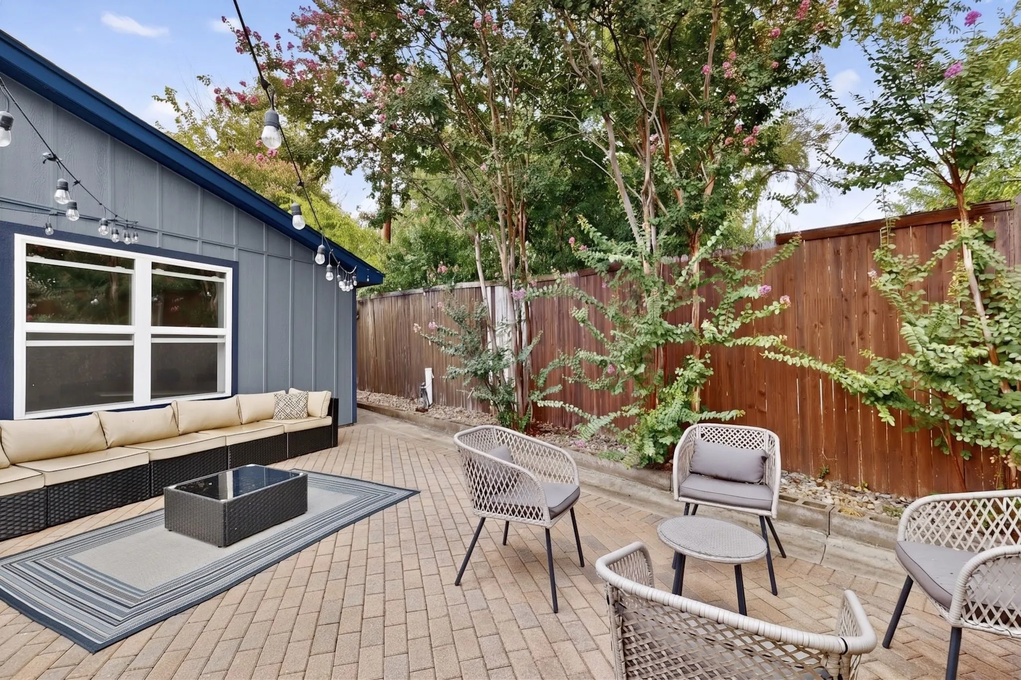 Paved brick patio area featuring a privacy fence, mature trees, and string lighting