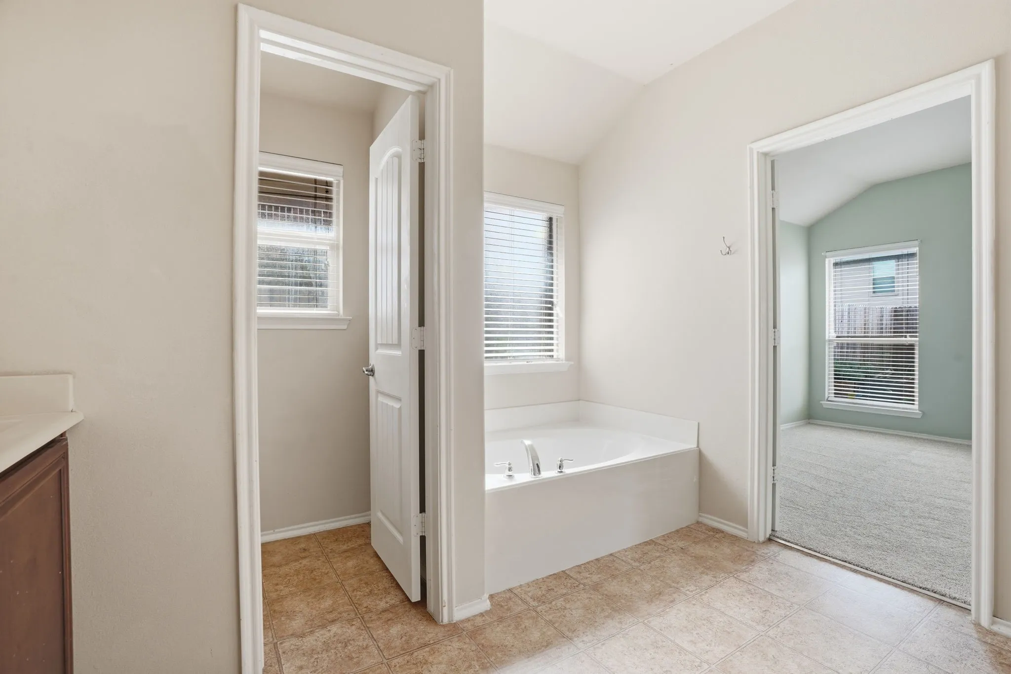 Built-in garden tub with chrome fixtures, two windows with horizontal blinds, neutral wall paint, and tile flooring