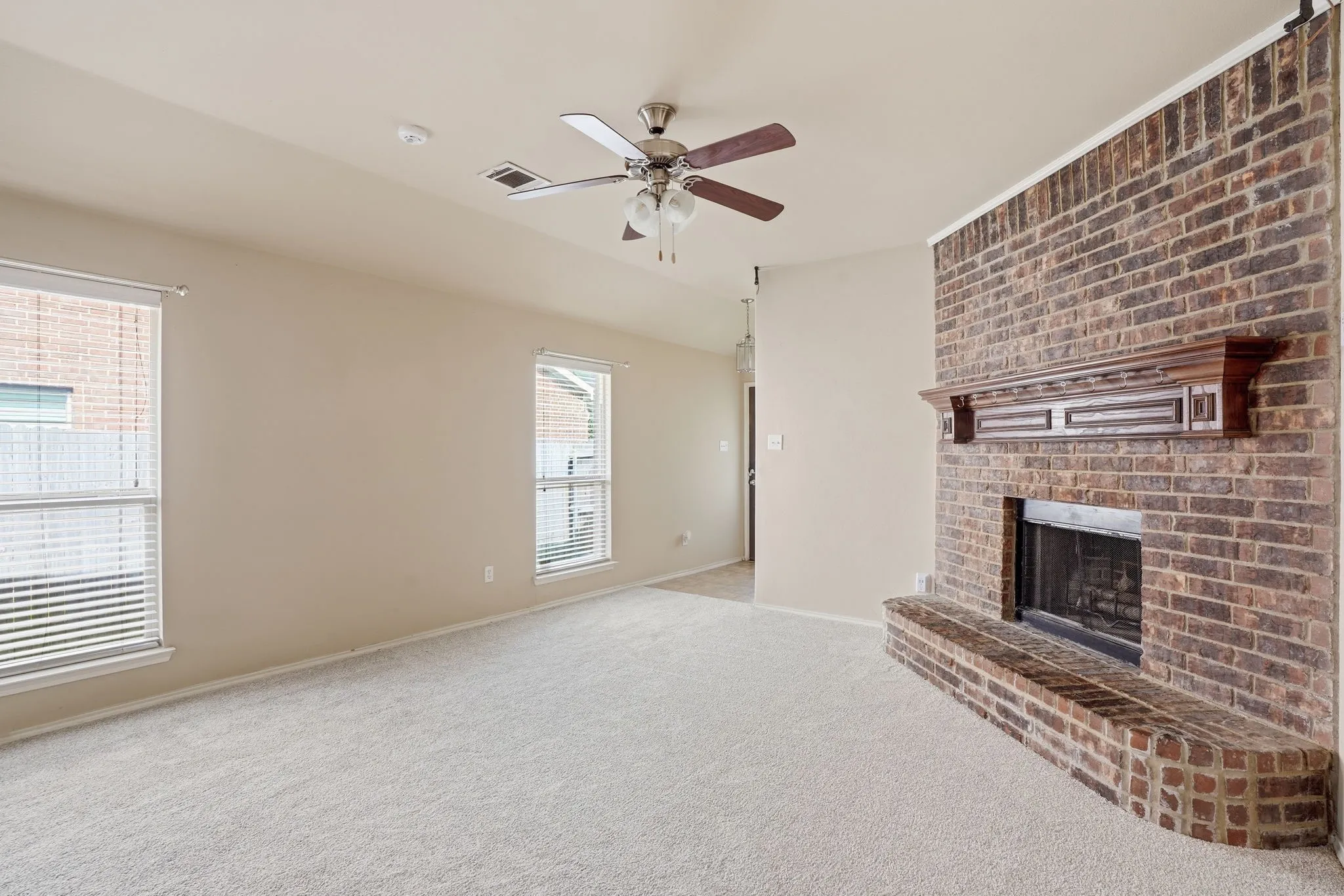 Spacious room featuring a brick fireplace with a carved wood mantel, light-colored carpet, and two windows with blinds