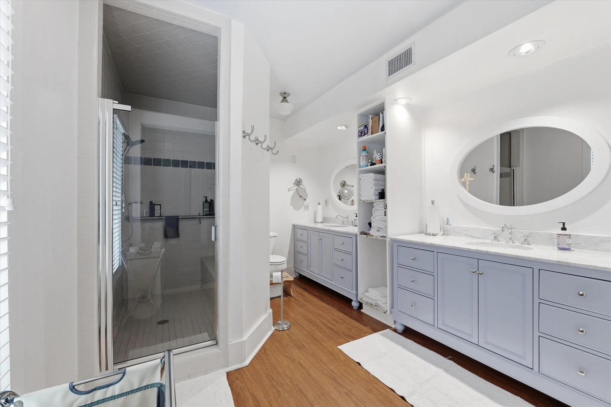 Bathroom featuring a glass-enclosed shower, dual vanity with light blue cabinetry, granite countertops, oval mirrors, and wood-finish flooring