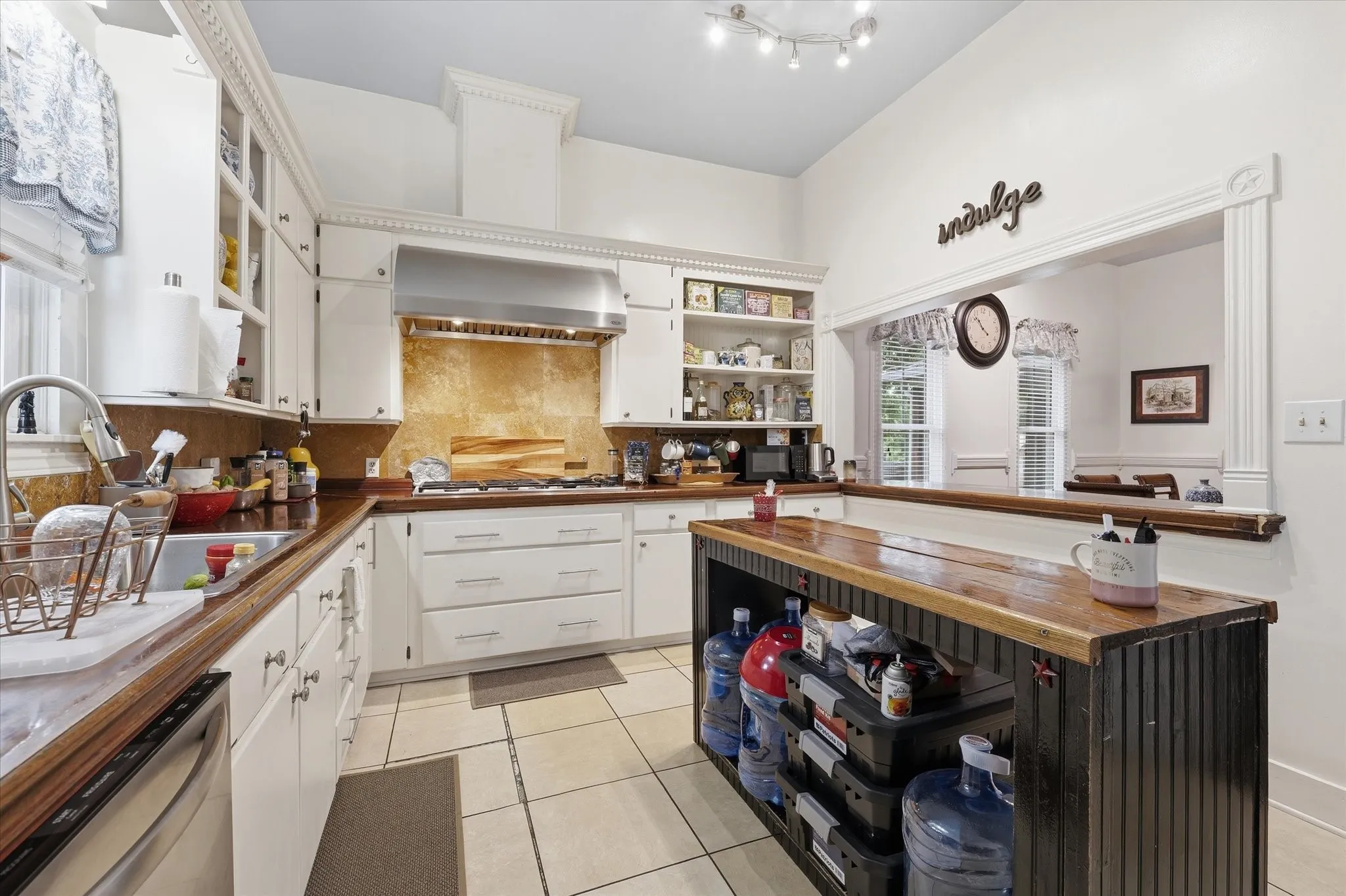 White cabinetry with crown molding, mahogany countertops, stainless steel range hood, tile flooring, and a pass-through window with a wood-finish bar top