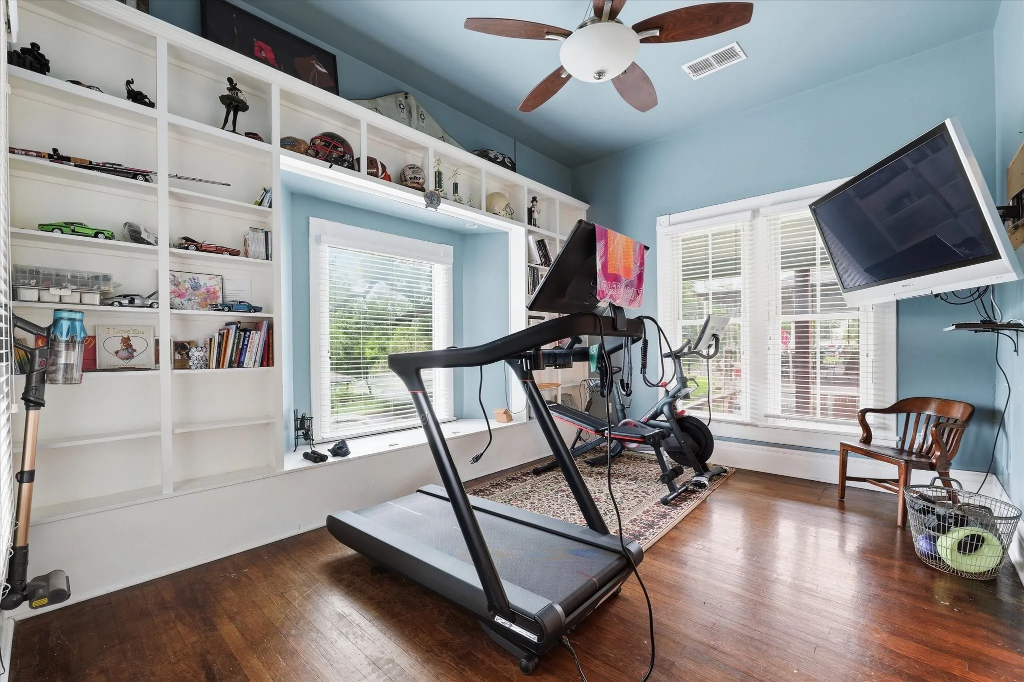 Hardwood flooring room featuring extensive built-in shelving, multiple windows with blinds, and a ceiling fan with integrated lighting