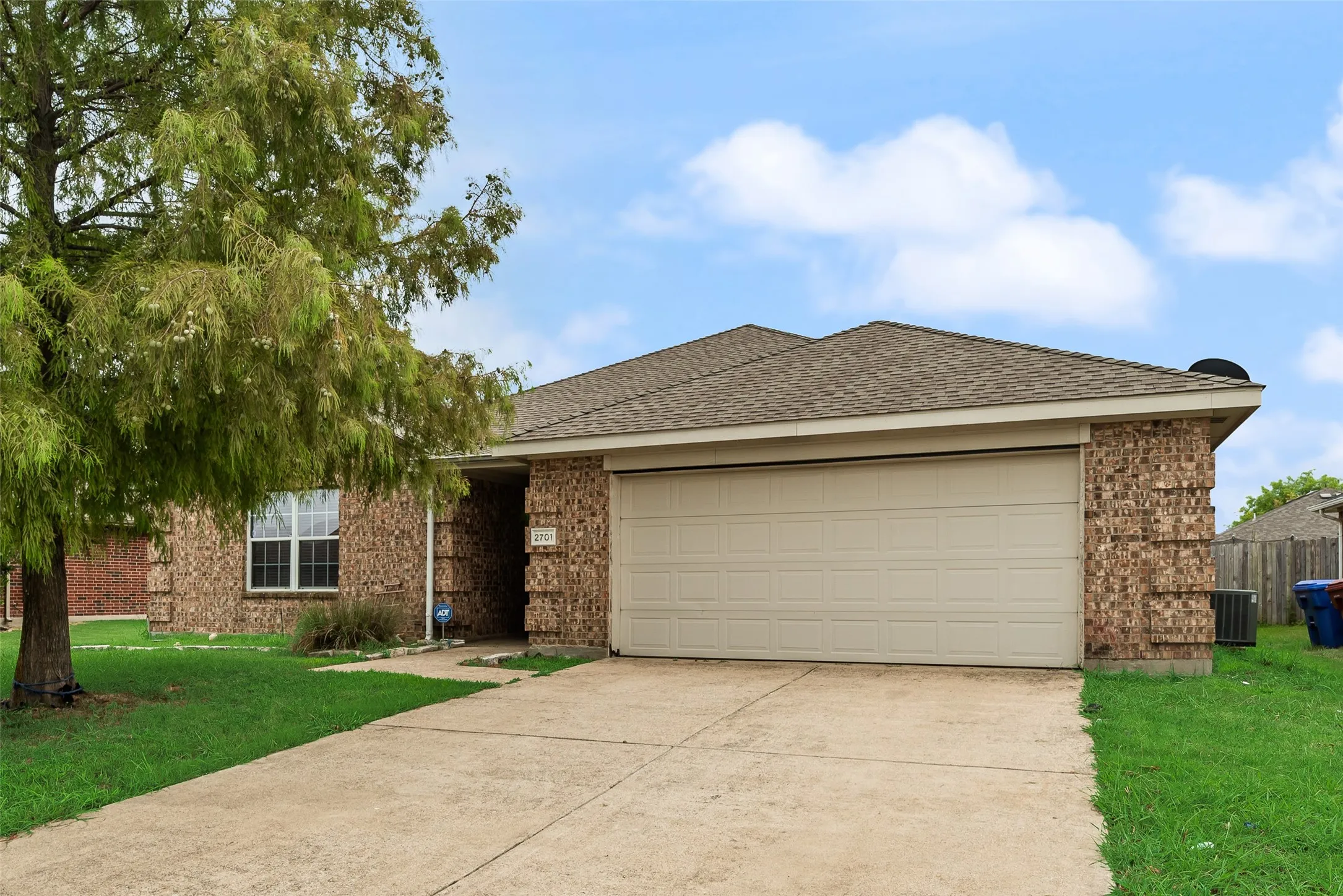 Brick facade home featuring an attached garage with a panel door, a concrete driveway, a composite shingle roof, a front lawn, and mature trees