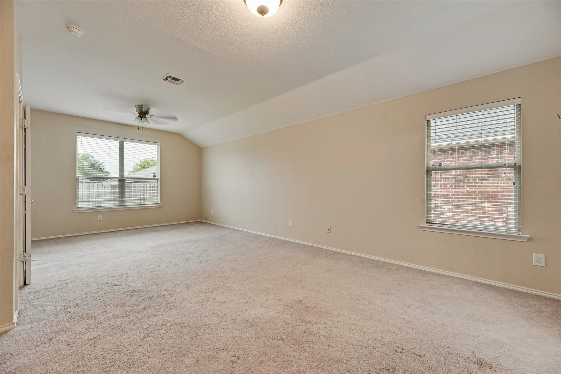 Spacious room featuring neutral carpet flooring, vaulted ceilings, and multiple windows with blinds