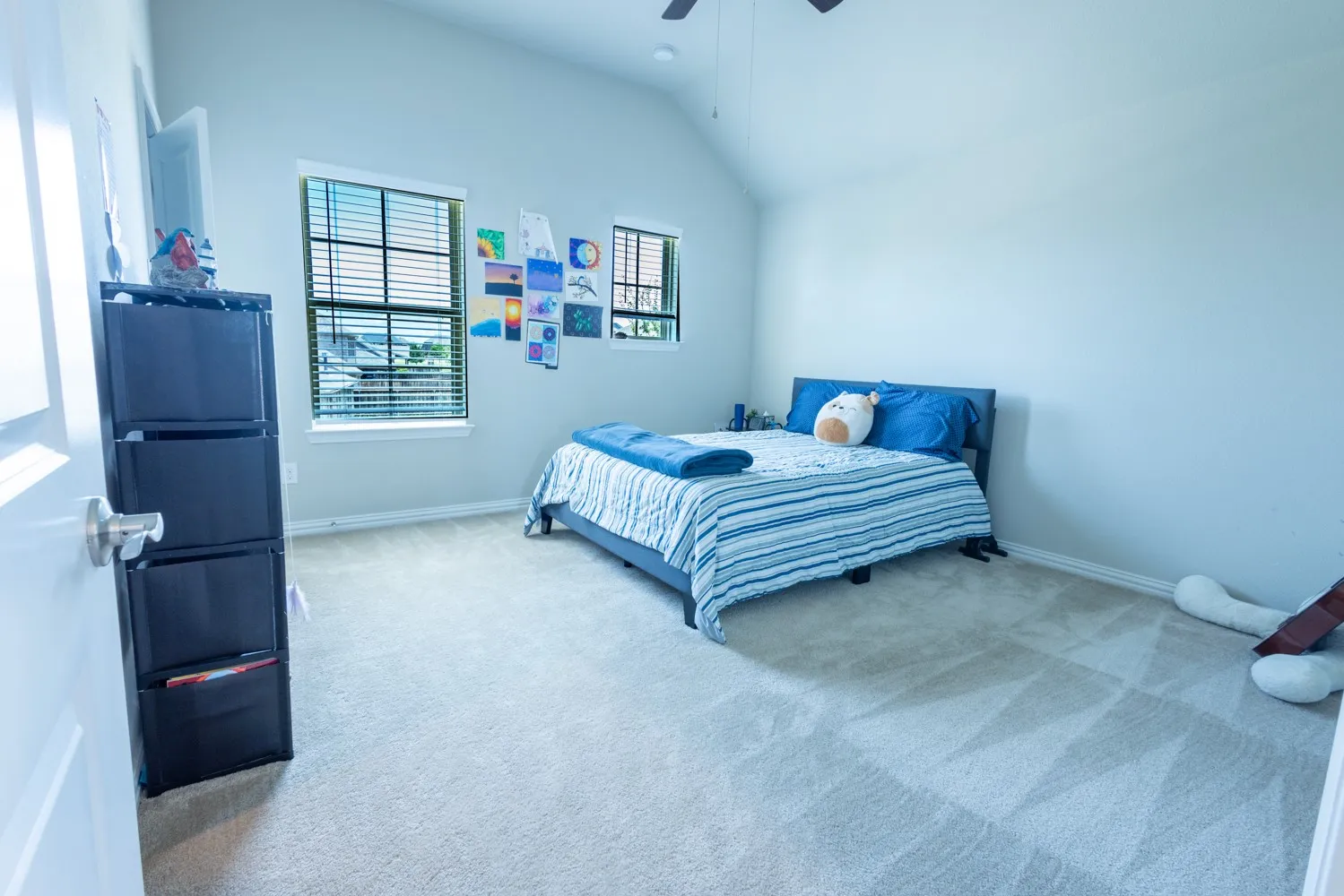 Room featuring vaulted ceilings, neutral-toned carpet flooring, two windows with horizontal blinds, and a ceiling fan